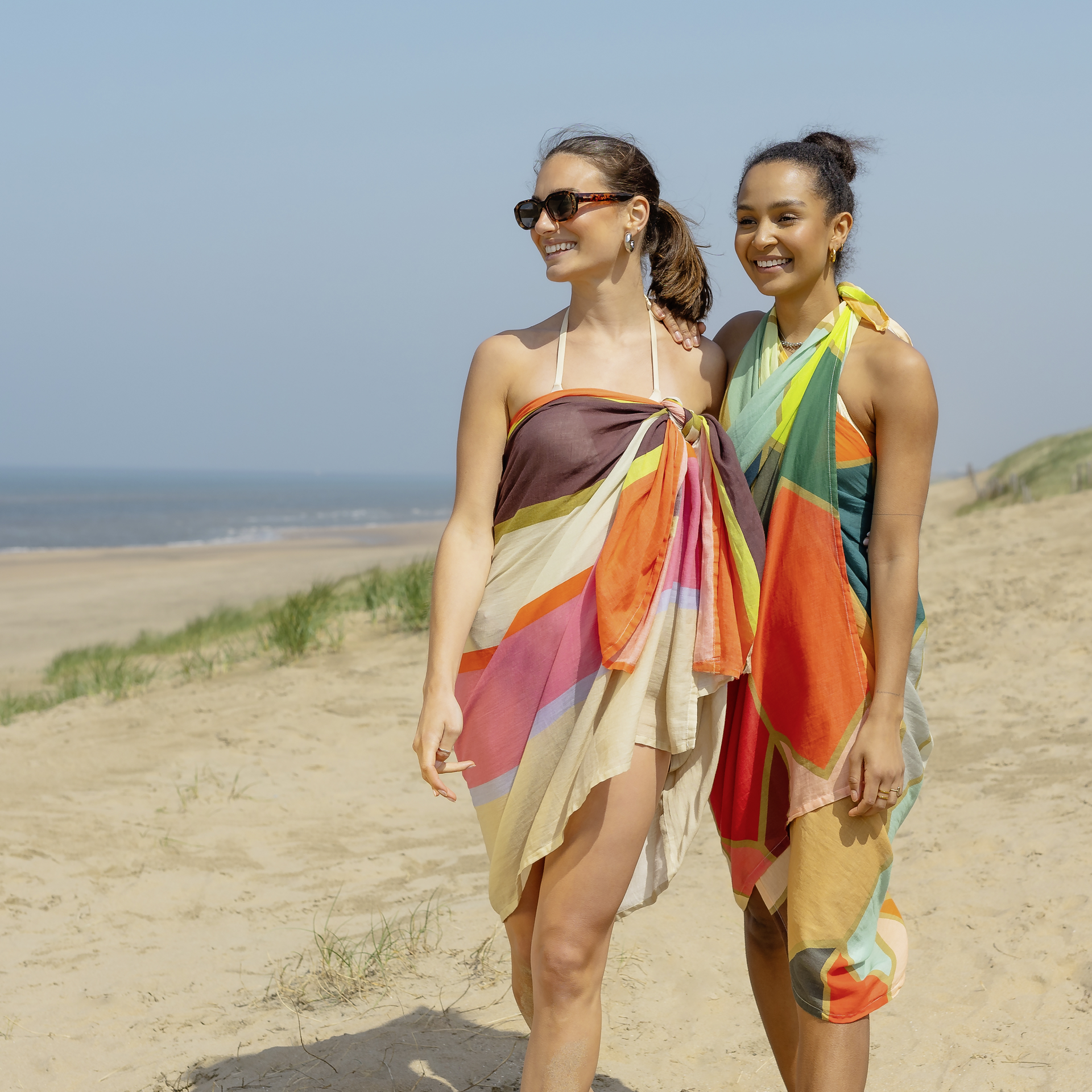 Zwei Frauen am Strand tragen farbenfrohe Pareos. Eine trägt Sonnenbrille. Im Hintergrund ist das Meer und ein Sandhügel zu sehen.