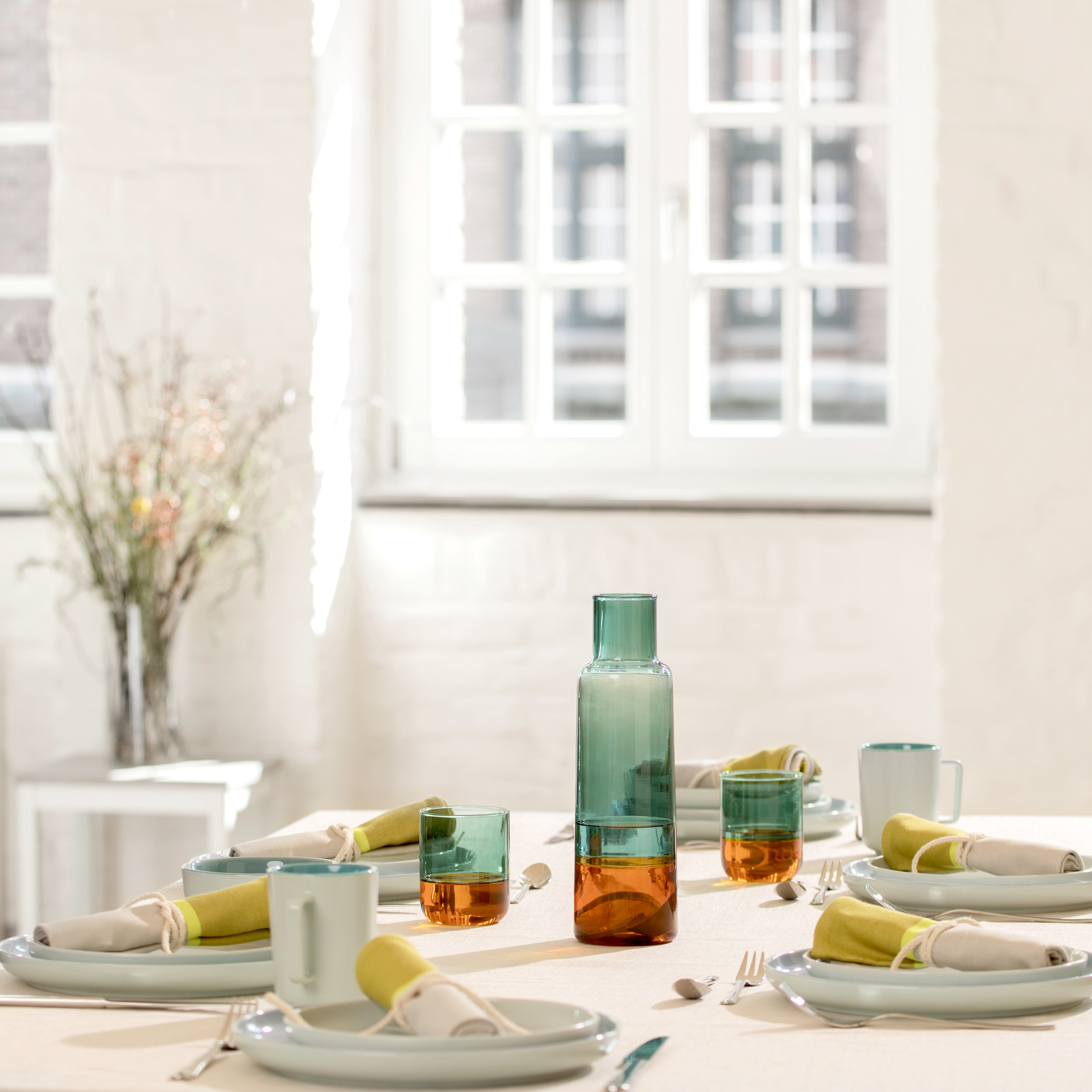 Ein gedeckter Tisch mit Tellern, Servietten, Besteck, Tassen und Gläsern 'amber' im 2er-Set. Eine Glaskaraffe ist mittig platziert. Helles Tageslicht im Hintergrund.