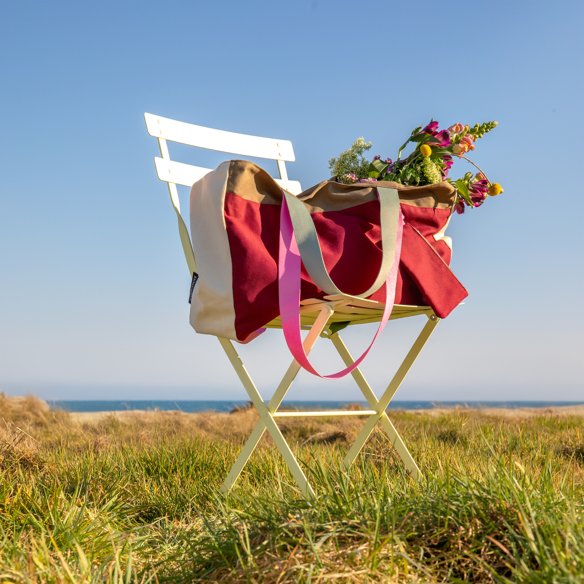 Draufsicht einer Canvas Tasche 'Emma' mit Blumen auf einem hellen Klappstuhl im Freien. Im Hintergrund sind Gras und ein blauer Himmel sichtbar. Die Tasche ist mehrfarbig mit pinken Trägern.
