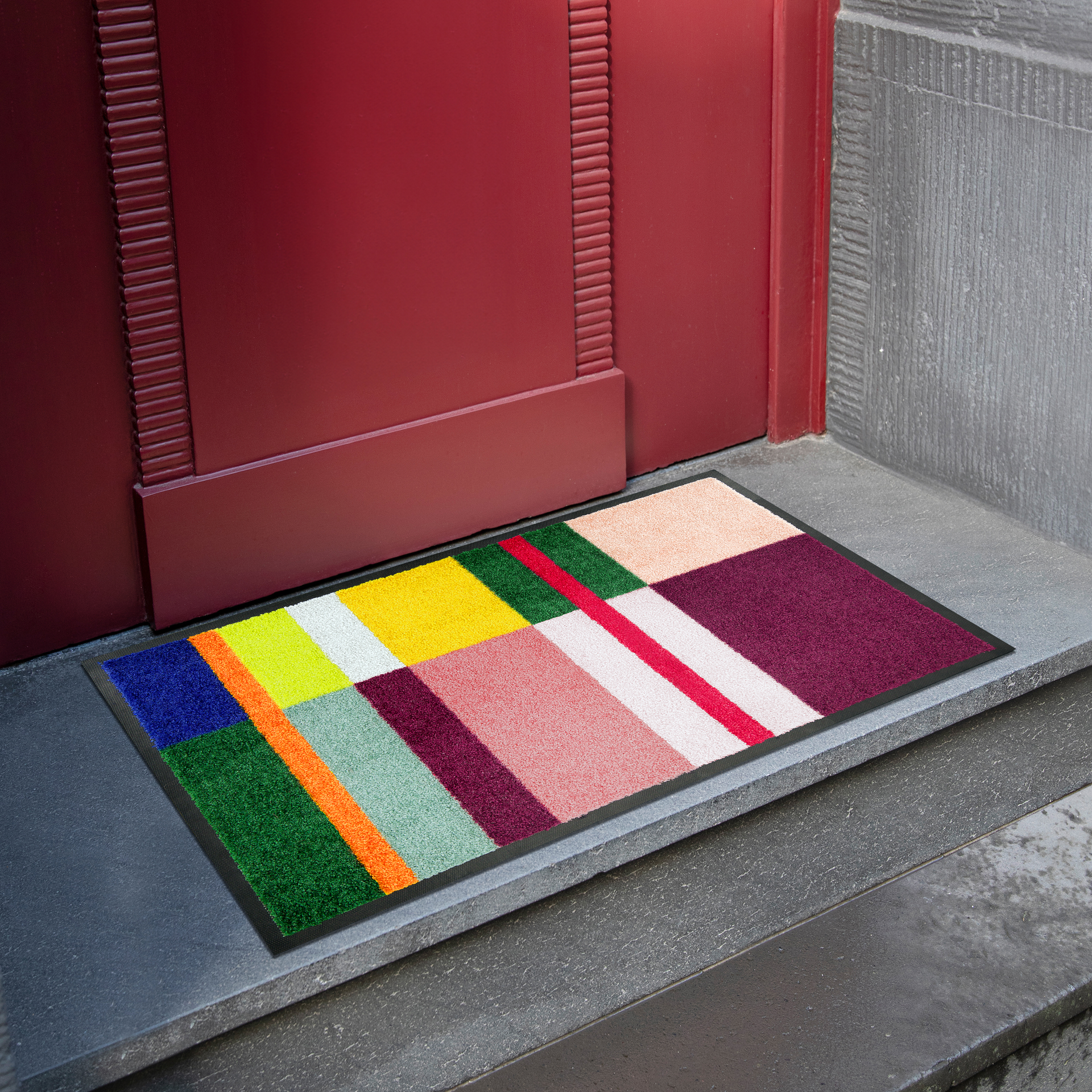 Ein Bodenbelag mit geometrischem Design in bunten Farben liegt vor einer weinroten Tür auf einer grauen Steintreppe.