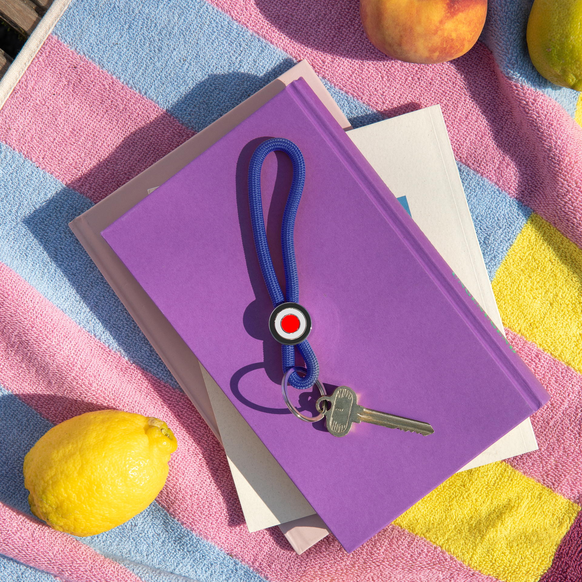 Blauer Schlüsselanhänger "Ringo deep blue" auf lila Buch. Daneben liegen ein Schlüssel, eine Zitrone und ein Pfirsich auf einem gestreiften Strandtuch.