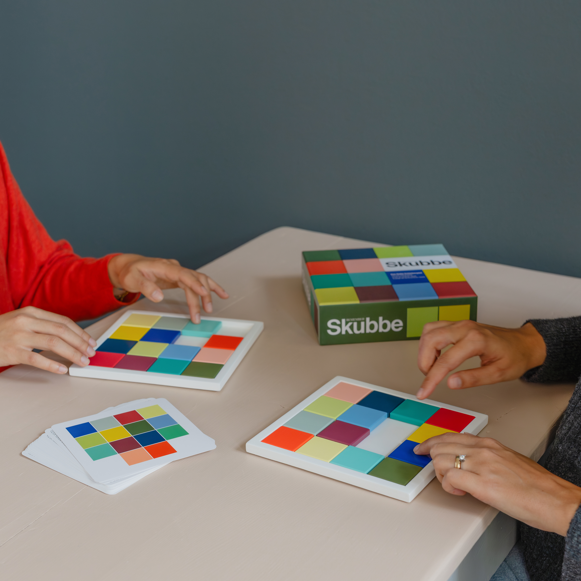 Zwei Personen spielen 'SKUBBE': Farbige Holzsteine in Spielbrett mit Vorlagekarten und Spielverpackung.
