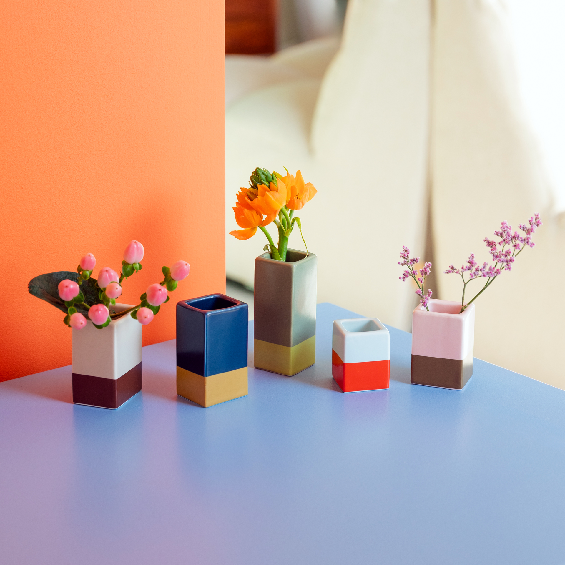 Cinque vasi colorati con fiori su un tavolo azzurro, di fronte a una parete arancione e a uno sfondo chiaro. I vasi sono di dimensioni diverse.