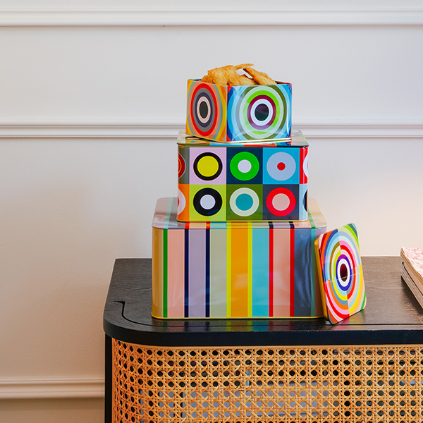 Trois boîtes de rangement rectangulaires colorées sur une table en bois noir, devant une façade en osier. En haut : boîte avec pâtisseries dorées, motif de cercles. Au milieu : carrés à cercles. En bas : bandes arc-en-ciel.