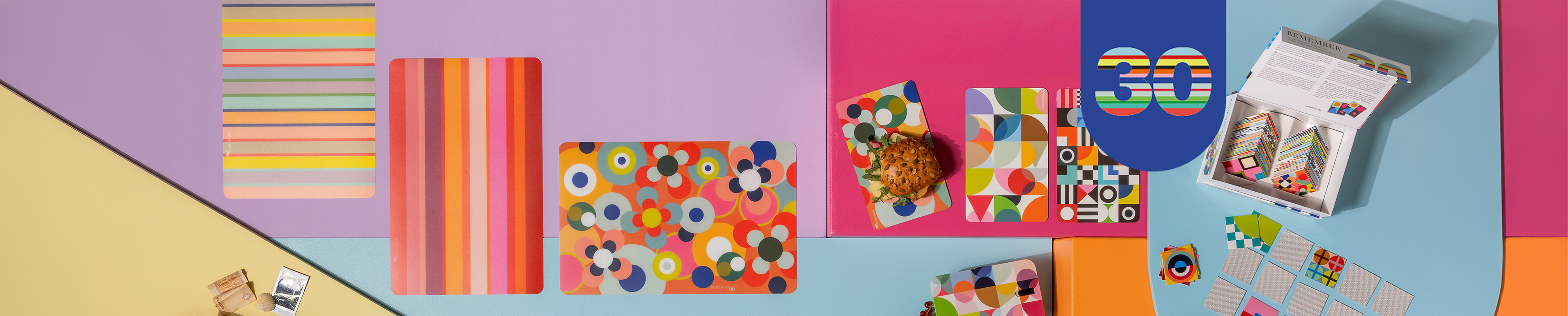 Composition multicolore et plate avec des blocs de couleur jaune, violet, rose et bleu. Des motifs sur des sous-verres et des cartes, en partie avec des aliments, complètent un jeu avec le chiffre 30.