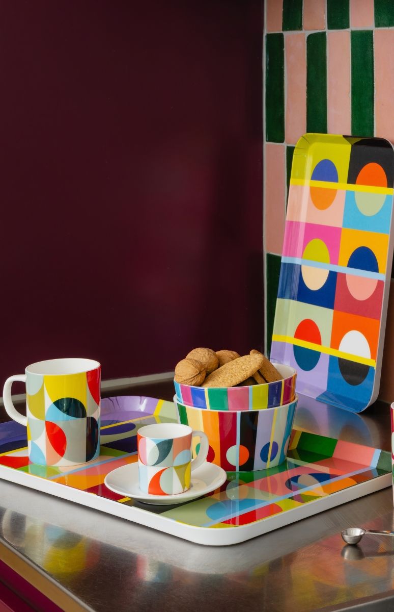 Bunt gemustertes Geschirr auf glänzender Arbeitsplatte: Tasse, Espressotasse mit Untertasse sowie zwei Schalen mit Keksen und Walnüssen auf rechteckigem Tablett. Dahinter lehnt ein Tablett an grün-rosa Kachelwand.