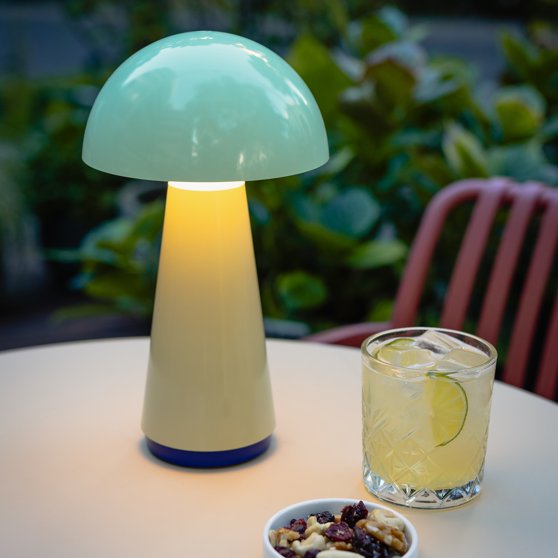 Mintfarbene Tischleuchte 'Bob' leuchtet auf einem Tisch, neben einem Cocktail und einer Schale mit Nüssen. Im Hintergrund sind grüne Pflanzen und ein Stuhl zu sehen.