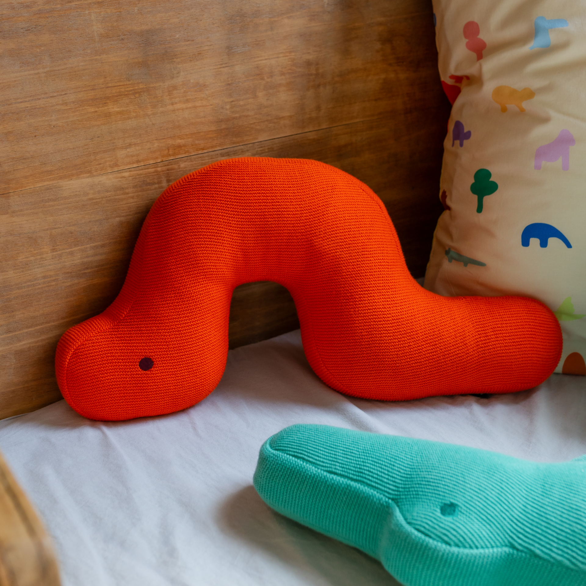 Orangefarbenes Kuschelkissen „Raupe Frieda“ in Form einer stilisierten Raupe mit blauem Punkt als Auge auf weißem Hintergrund.