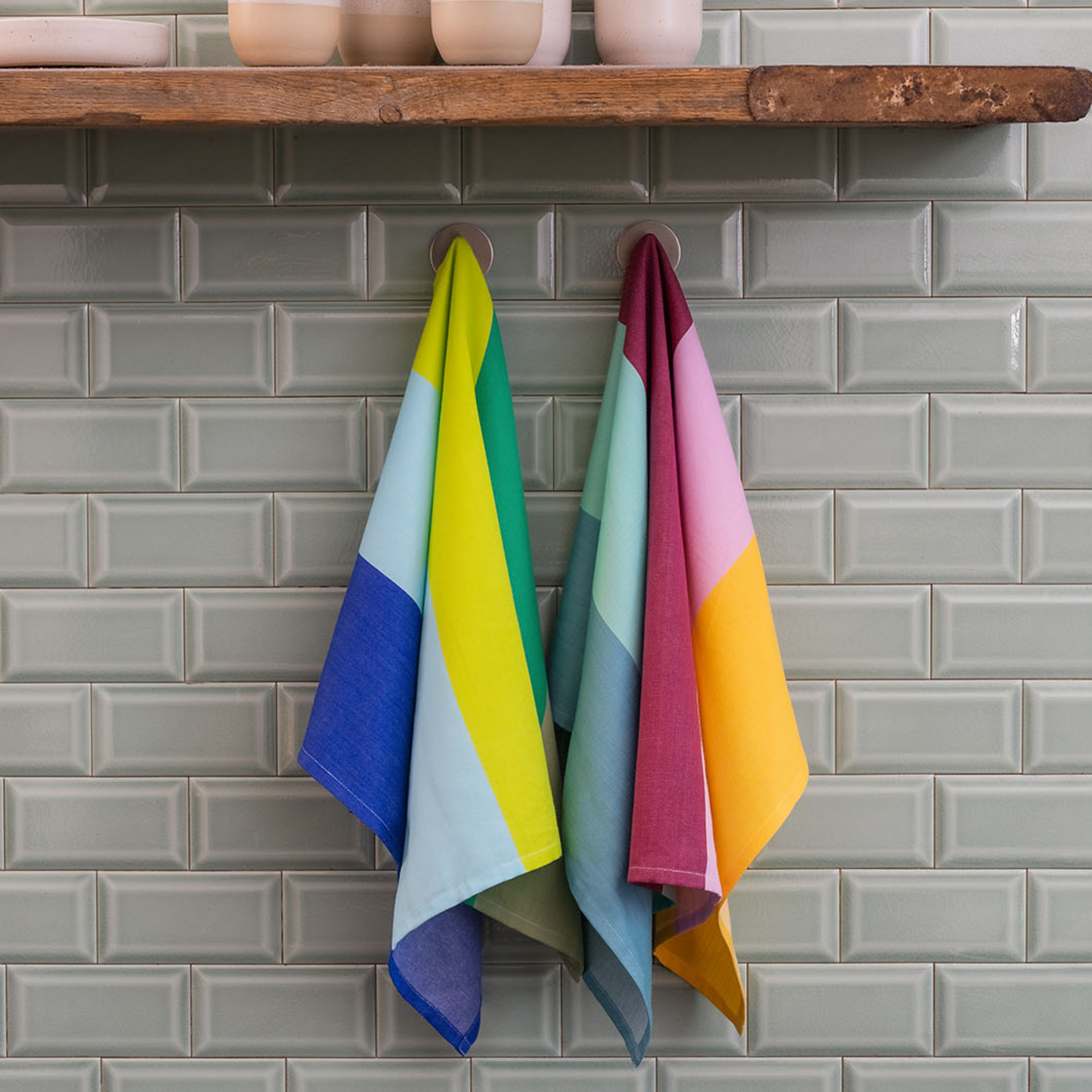 Zwei bunte Trockentücher-Sets hängen an Haken vor einer hellgrünen Kachelwand. Über den Tüchern befindet sich ein Holzregal mit Geschirr.