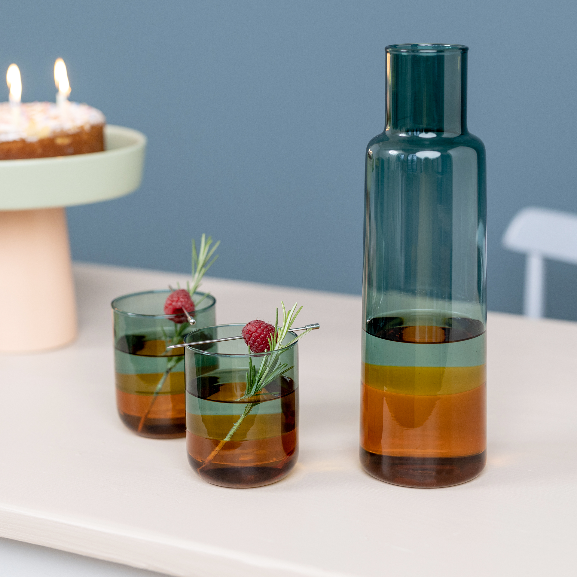 Zwei 'amber' Gläser mit Getränk, dekoriert mit Himbeere und Rosmarin, neben einer passenden Karaffe auf einem Tisch. Im Hintergrund ein Kuchen mit Kerzen.