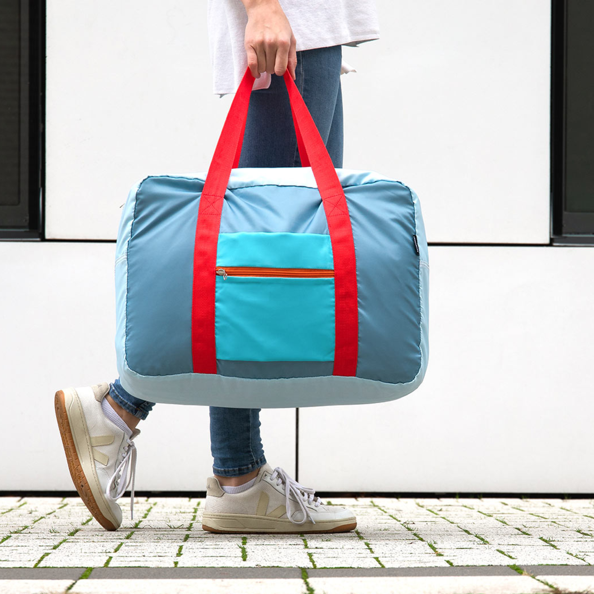 Eine Person trägt die hellblaue 'Tom' Reisetasche an roten Trägern. Die Tasche hat eine blaue Außentasche mit Reißverschluss. Der Hintergrund ist eine helle Wand.