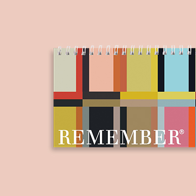 Tischkalender mit farbigen geometrischen Mustern und Spiralbindung, beschriftet mit dem Wort "REMEMBER" auf rosa Hintergrund.