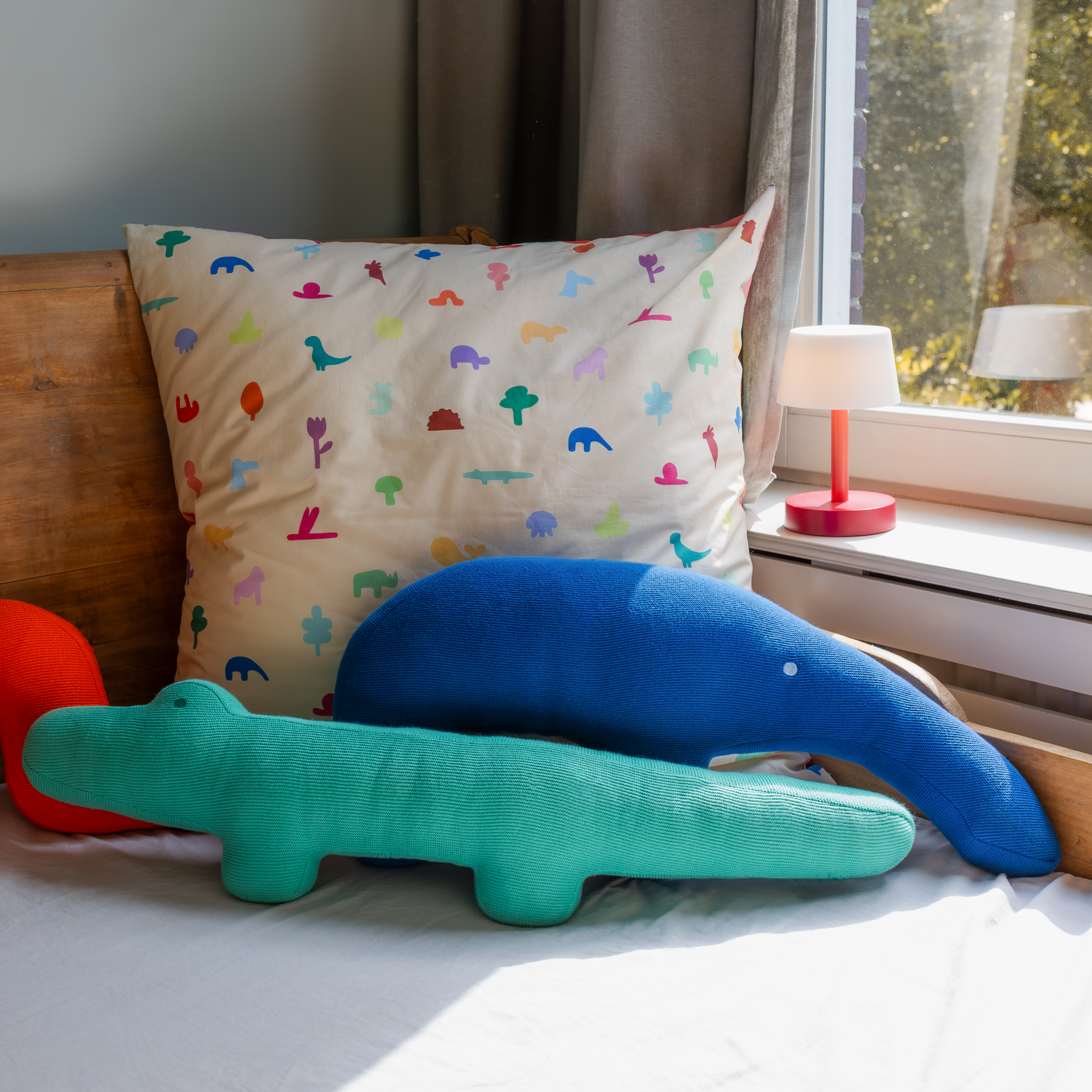 Kuschelkissen: Ein Kissen mit bunten Tiermotiven, davor ein blaues Ameisenbär-Kissen und ein grünes Krokodil-Kissen auf einem Bett.