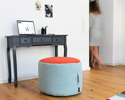 Ein blauer Pouf mit orangem Sitz steht vor einem Schreibtisch mit Laptop. Im Hintergrund ist eine Person undeutlich in einer Türöffnung zu sehen.