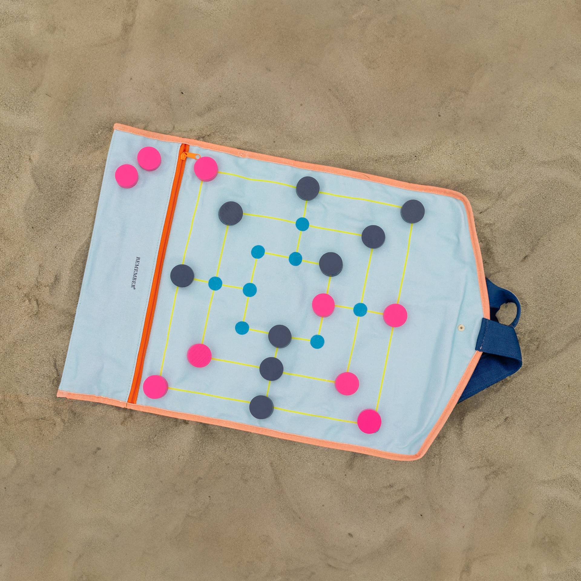 Textiles 'Roll-Up' Brettspiel auf Sand. Das aufgeklappte Spiel zeigt ein Mühle-Spielbrett mit bunten Spielsteinen und einer Reißverschlusstasche.