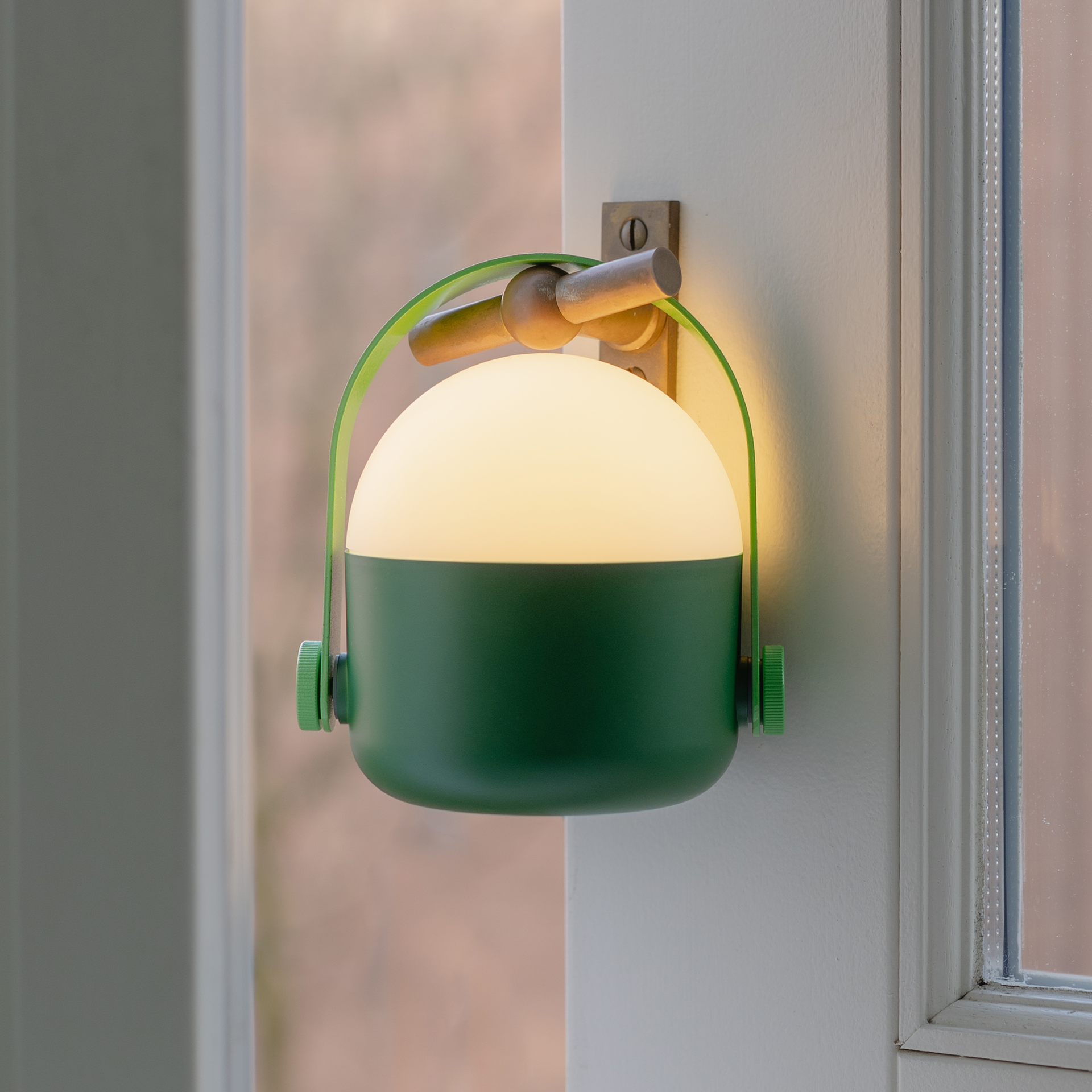 Eine grüne Leuchte mit Henkel und warmweißem Licht ist an einer Wand neben einem Fenster montiert.