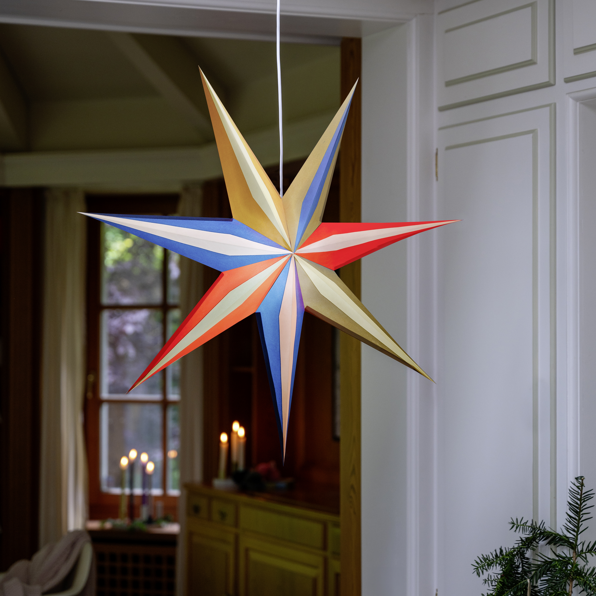 Une étoile en papier multicolore 'Mago' de différentes couleurs est suspendue dans la pièce. La lumière brille à travers une fenêtre à l'arrière-plan.