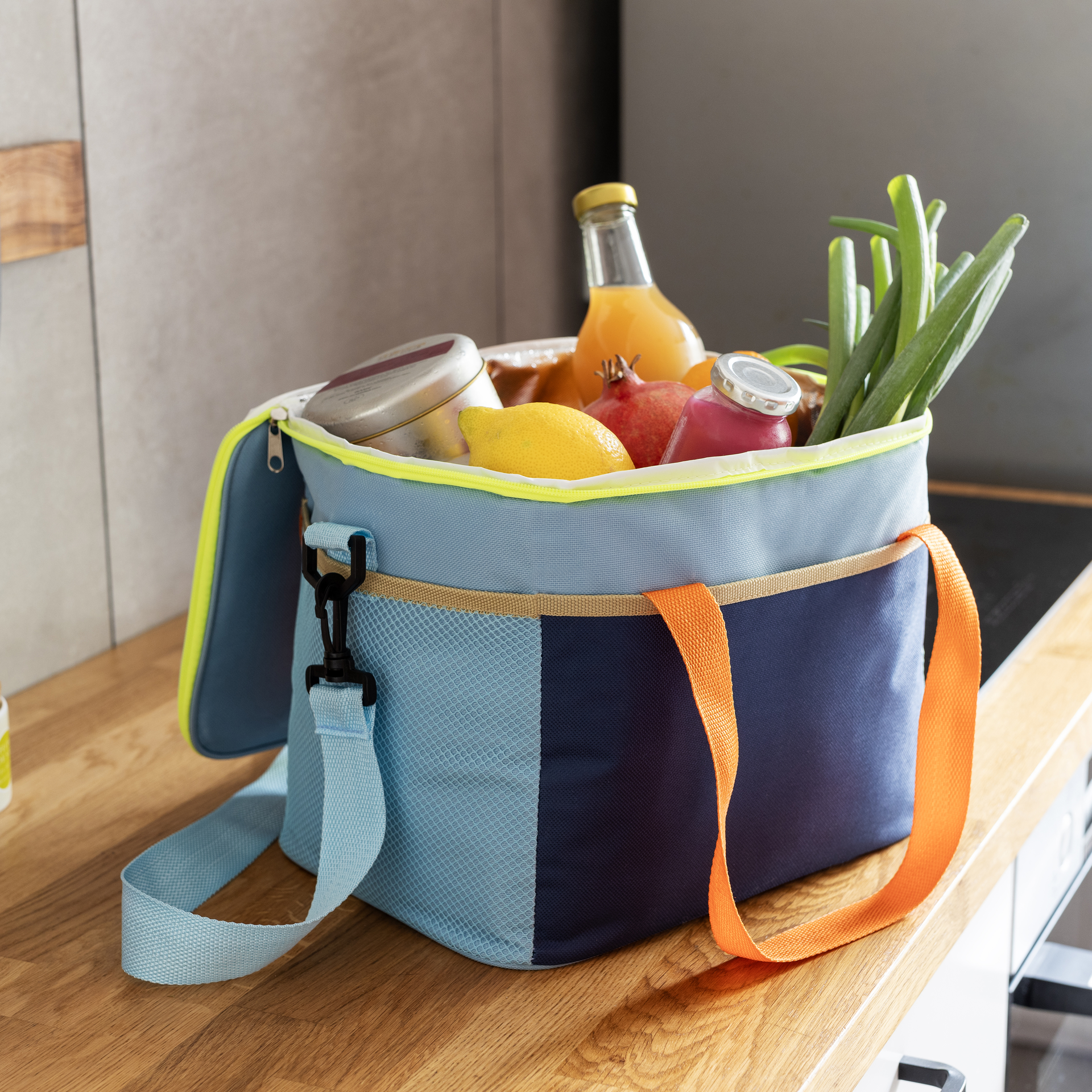 Geöffnete, hellblau-dunkelblaue Kühltasche 'Nick' mit Tragegurt und orangefarbenen Henkeln steht auf einer Küchentheke. Sie ist gefüllt mit Getränken, Zitrone und Frühlingszwiebeln.
