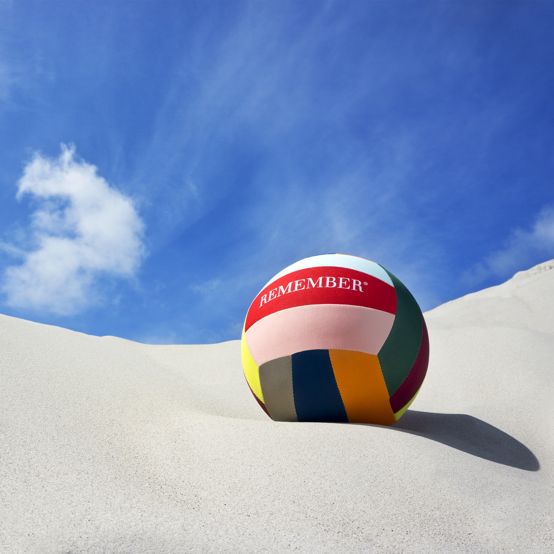 Ein farbenfroher Freizeitball mit der Aufschrift "REMEMBER" liegt im Sand unter blauem Himmel mit Wolken.