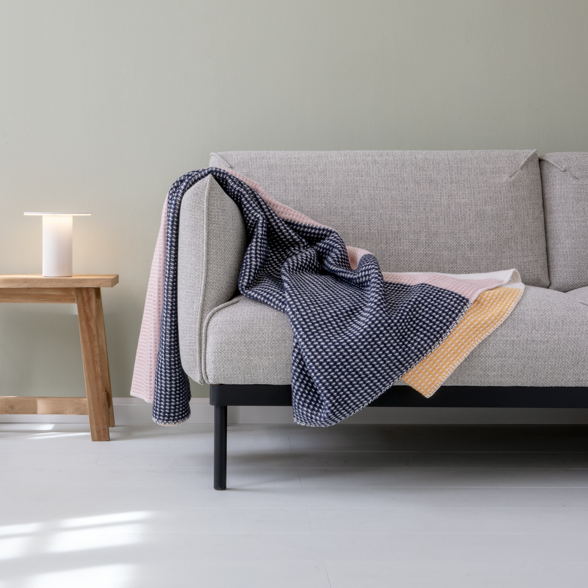Eine Gala-Decke mit Waffelmuster in Blau, Rosa und Gelb liegt auf einer grauen Couch. Ein Holzhocker mit Lampe steht daneben.