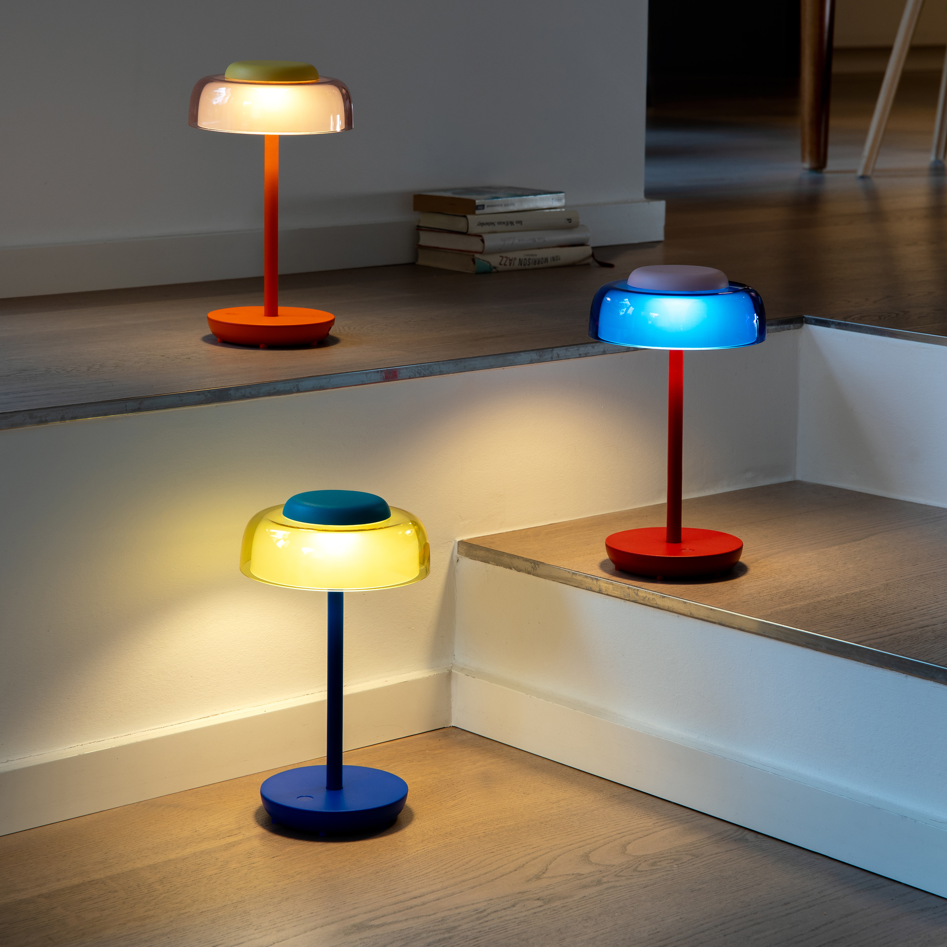 Tre lampade da tavolo colorate (modello Juno) sono su gradini di legno in una stanza. Una blu con paralume giallo emana luce calda, una arancione con paralume trasparente luce intensa, una rossa con paralume blu luce blu. Libri sullo sfondo.