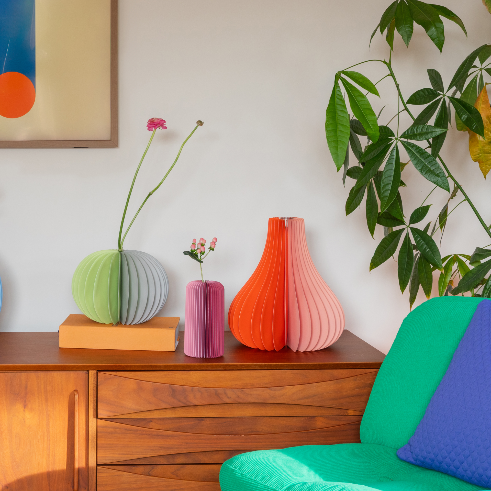 Auf einer Kommode stehen bunte Vasen, eine mit Blumen. Rechts eine Zimmerpflanze, und ein farbenfroher Sessel. An der Wand hängt ein Bild mit geometrischen Formen.