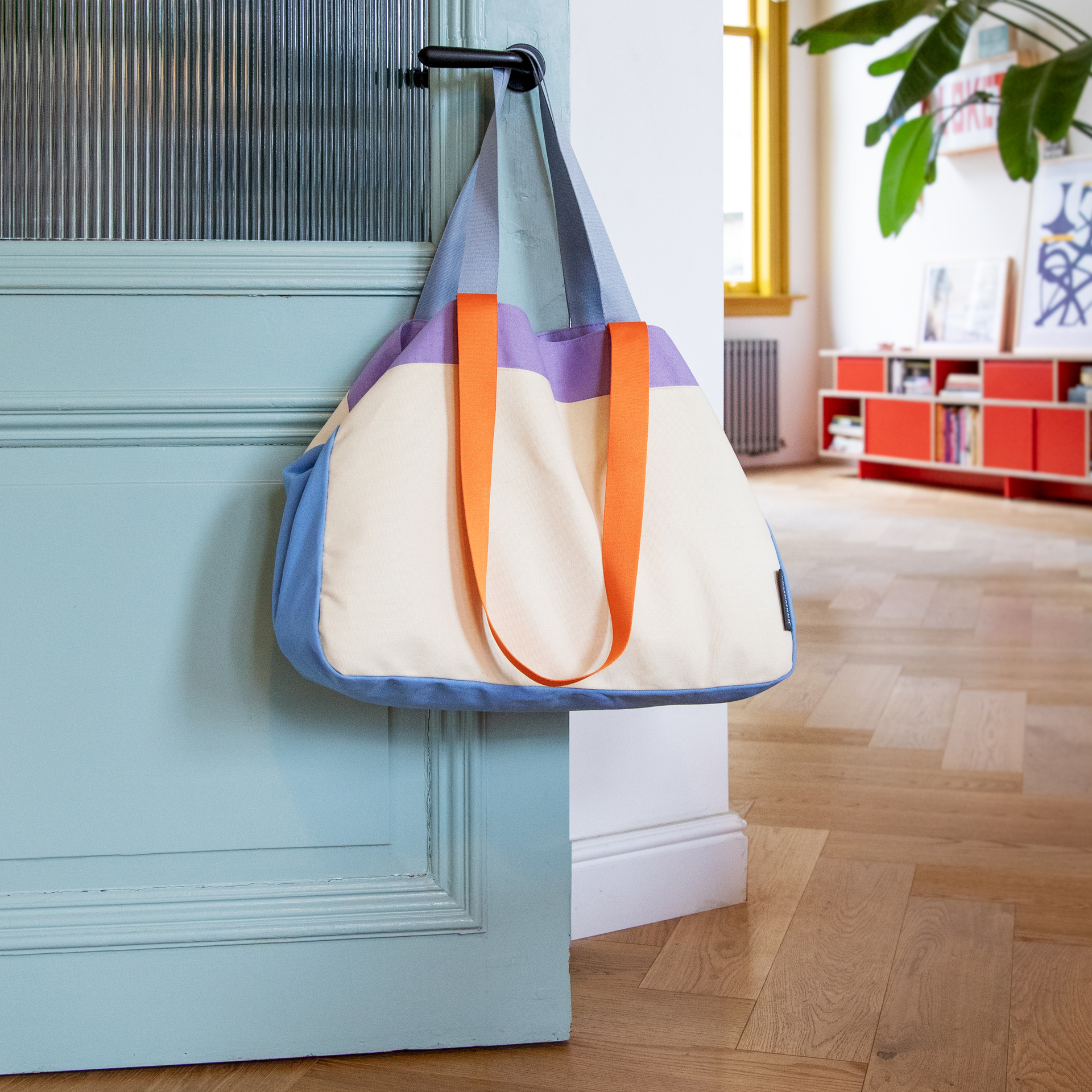 Canvas Tasche 'Toni' in blau, creme, lila mit orangefarbenen Trägern, hängt an einer türkisfarbenen Tür. Im Hintergrund ein Raum mit rotem Regal.