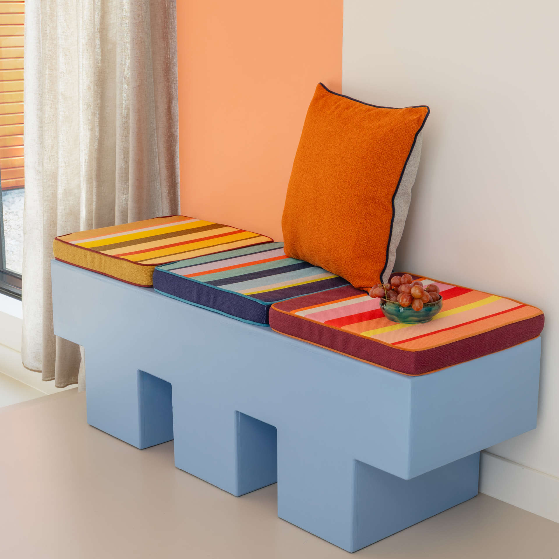 Blauer Hocker mit bunten Kissen und orangem Kissen an der Wand. Eine Schale mit Weintrauben steht auf einem der Kissen. Im Hintergrund Gardinen und eine farbige Wand.