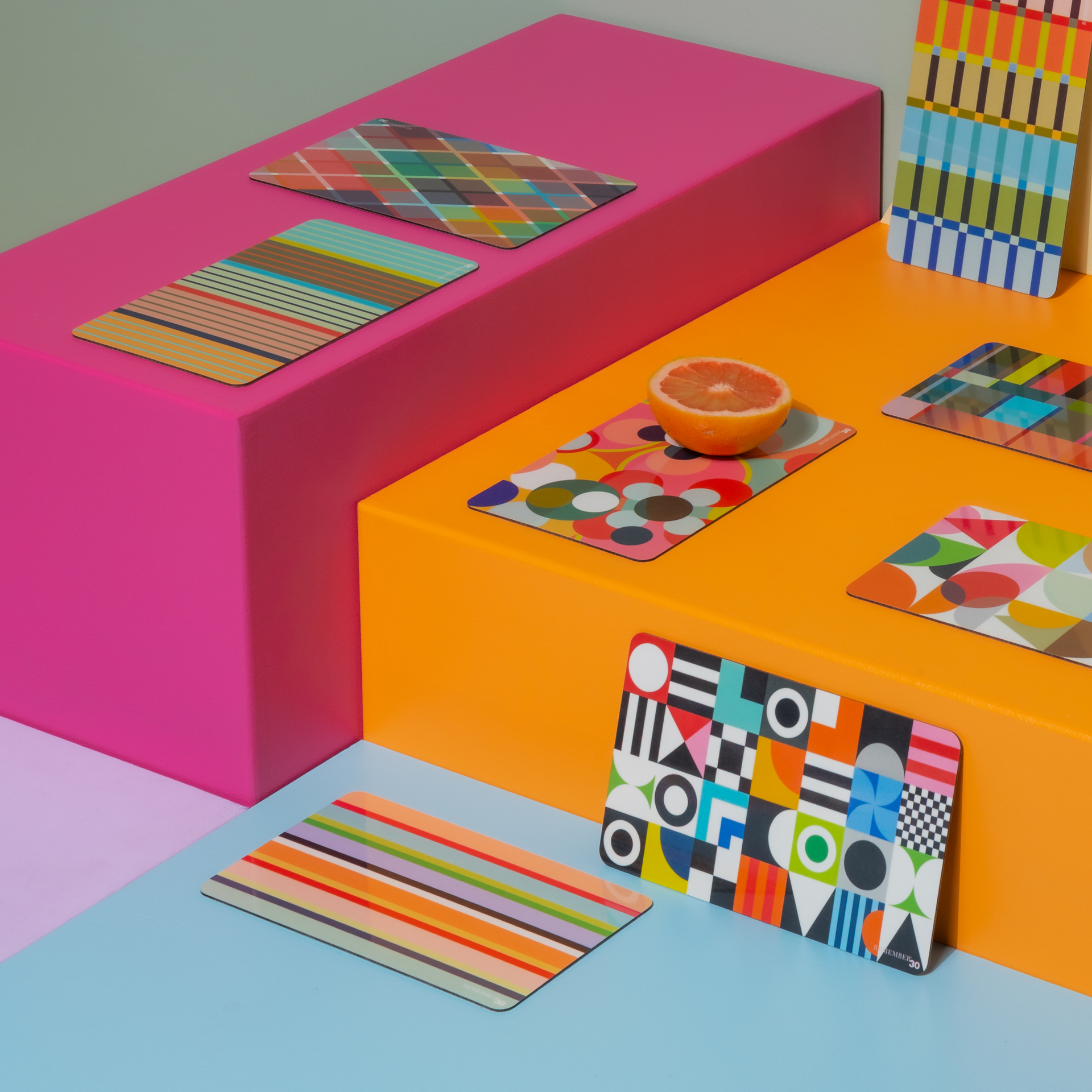Mehrfarbiges Set: Geometrische Muster, Streifen und Punkte auf Platzsets, arrangiert auf pinkfarbenen, orangen und blauen Untergründen. Eine halbe Orange ergänzt das Ensemble.