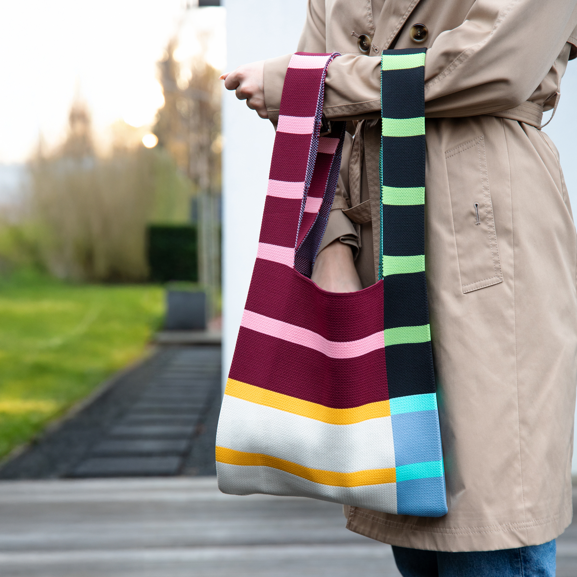Ein Model in Trenchcoat präsentiert die farbenfrohe Stricktasche 'Scala' mit Streifen in Rosa, Lila, Gelb, Weiß, Schwarz und Blau.