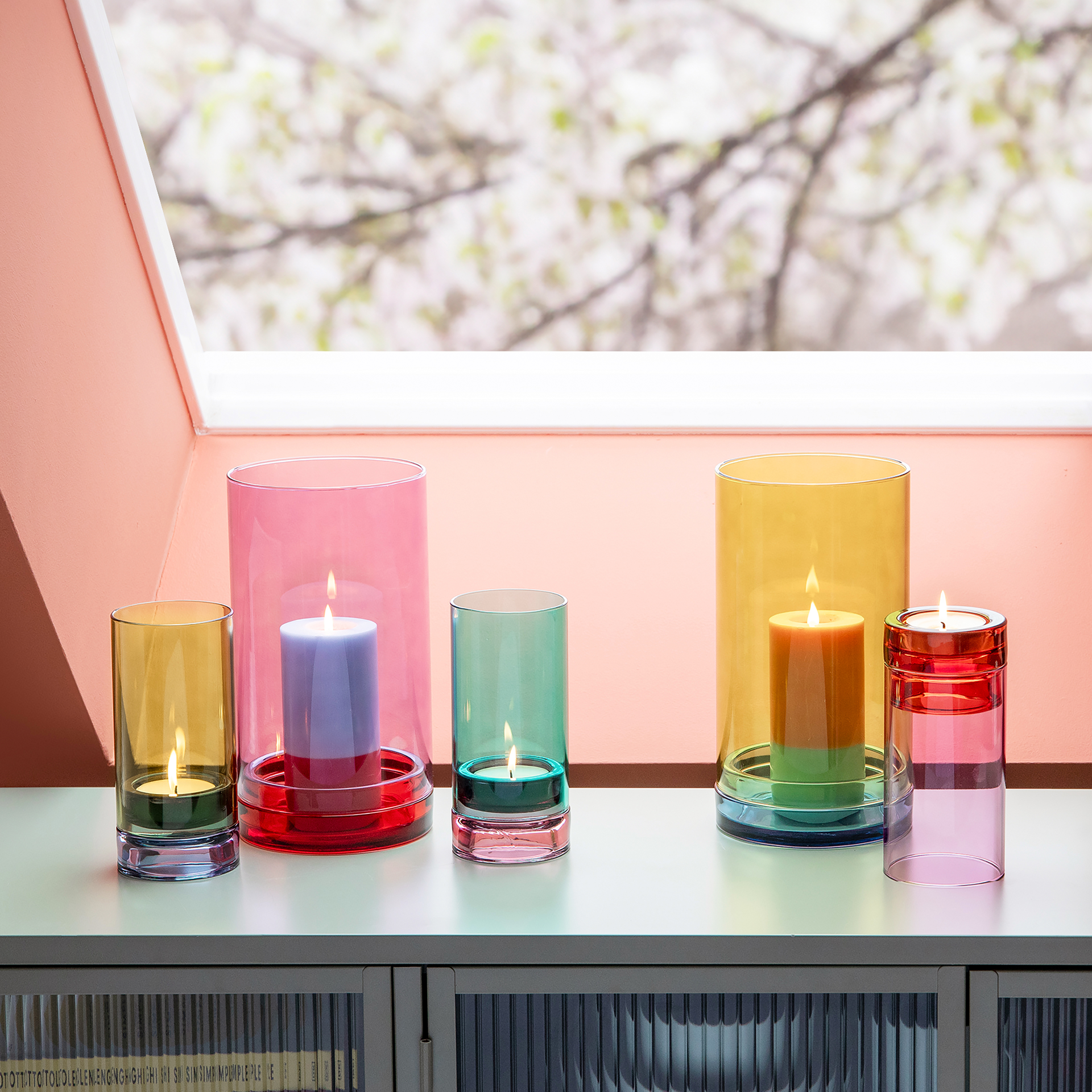 Fünf farbige Windlichter 'Lys' aus Glas mit brennenden Kerzen auf einer Ablage vor einem Fenster. Die Farben sind Aqua, Gelb, Rot und Pink.