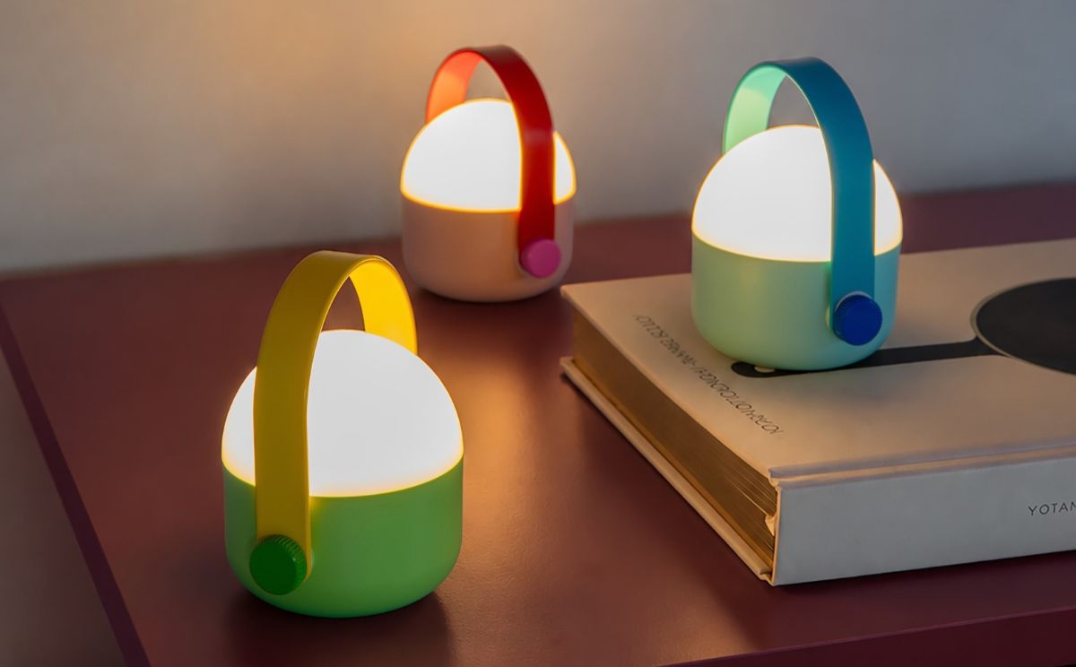 Trois lanternes modernes lumineuses sur une table rougeâtre. Devant : socle vert, poignée jaune, bouton rotatif vert. Derrière : socle rose clair, poignée rouge. Arrière droit : socle bleu clair, poignée bleue. Partiellement masquées par le livre.