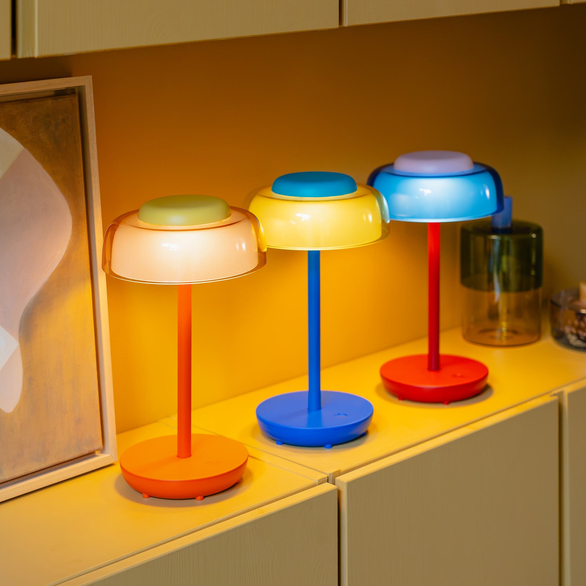 Tre lampade da tavolo Juno in diversi colori vivaci su uno scaffale. I piedini colorati e i paralumi luminosi risplendono in calde tonalità di giallo e in un fresco blu. Il set mette in risalto la varietà della serie Juno senza fili.