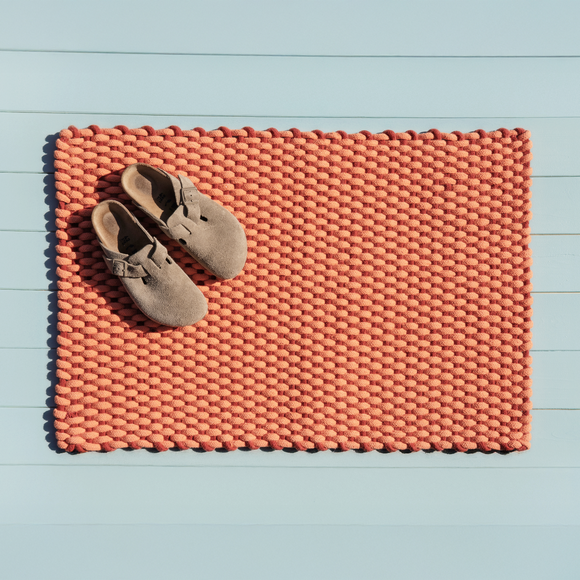 Handgewebte, korallenfarbene Fußmatte mit Struktur auf blauem Untergrund. Ein Paar Schuhe liegt auf der linken Seite der Matte.