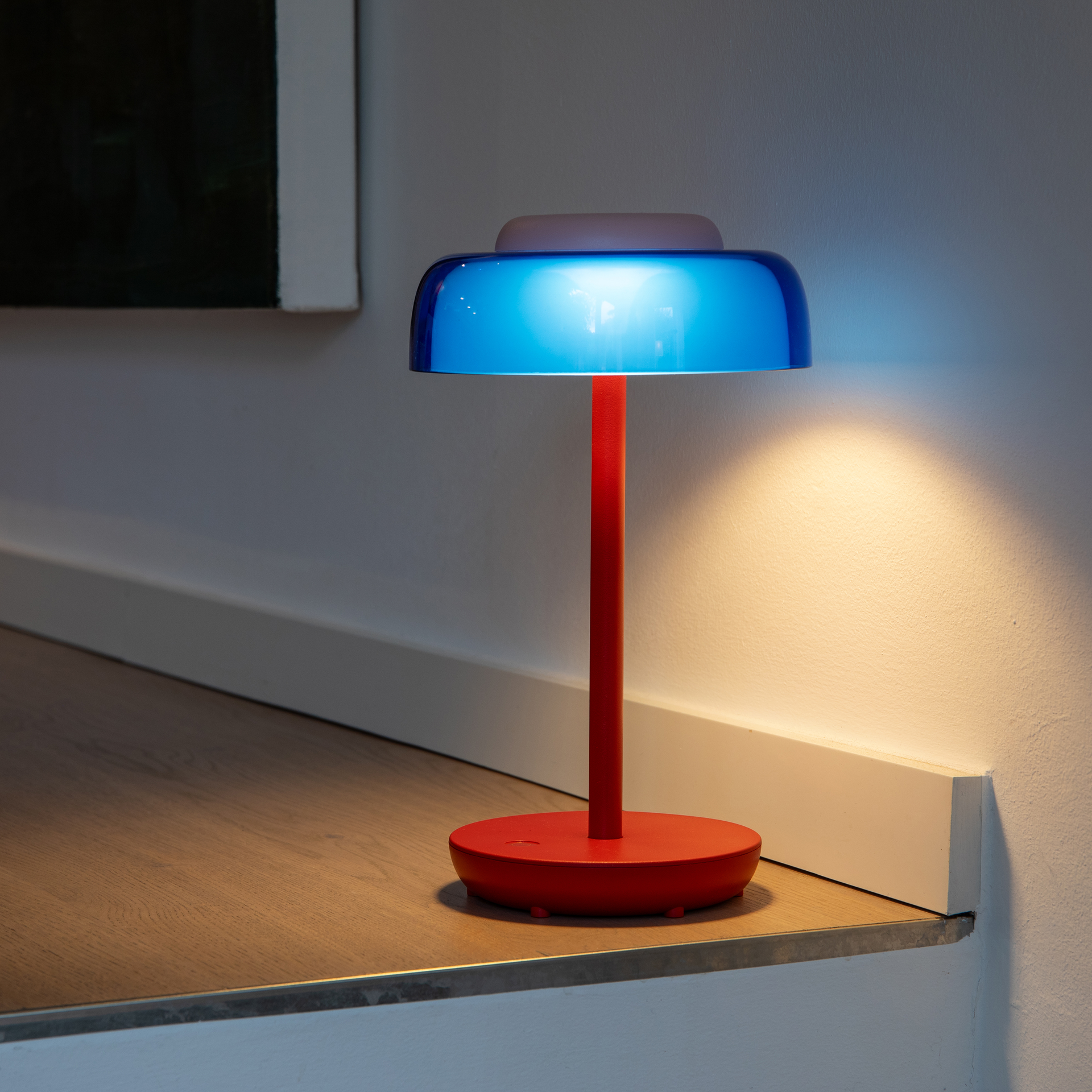 Lampada da tavolo senza fili Juno, con base e stelo in metallo rosso dalle linee slanciate. Paralume in plastica trasparente blu con comando touch rosa per regolare la luminosità. Sulla base il simbolo accensione/spegnimento.