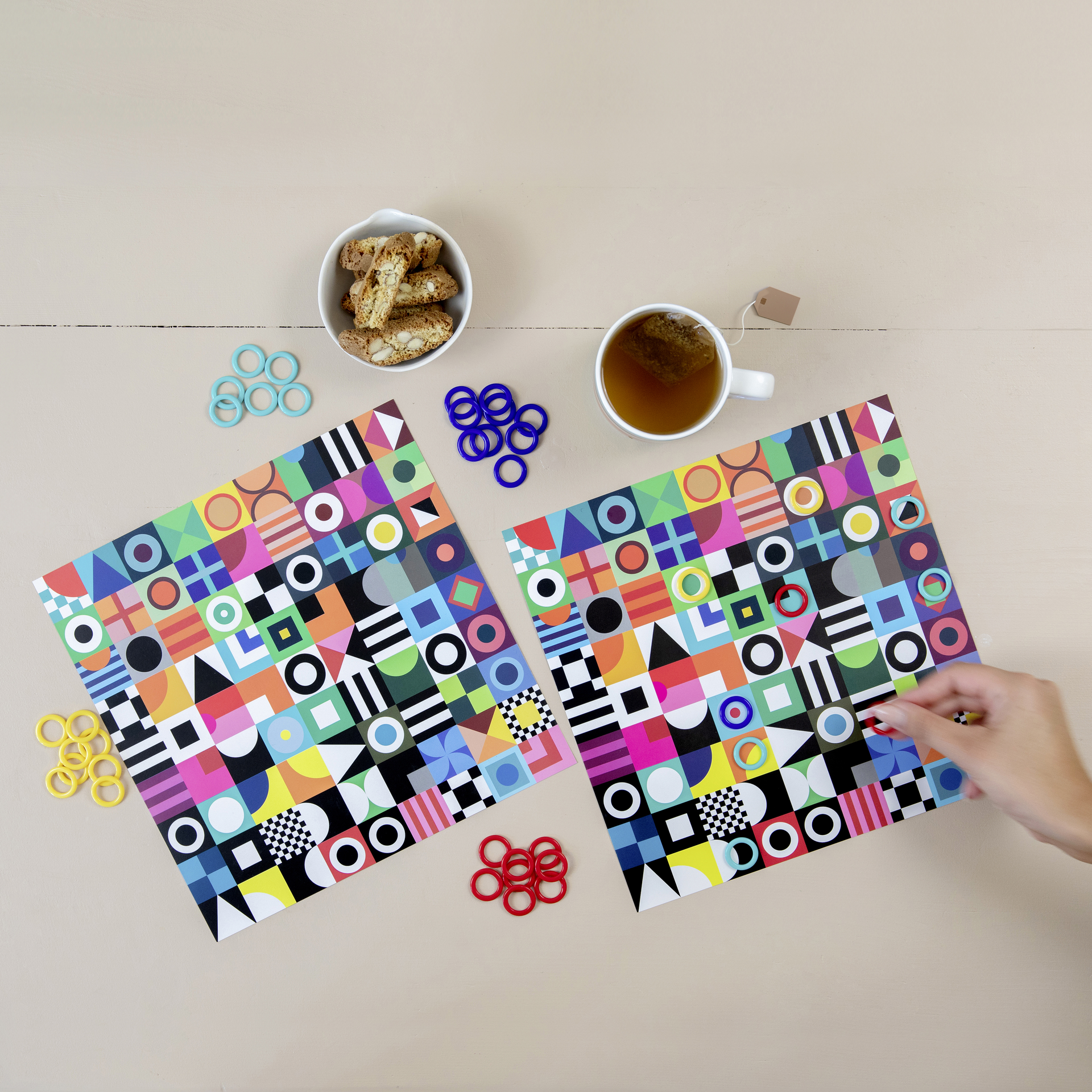 Zwei Spielblätter des Double Suchspiels mit geometrischen Mustern, farbigen Spielringen, Gebäck und einer Tasse Tee auf beigem Untergrund. Eine Hand platziert einen roten Ring auf ein Blatt.
