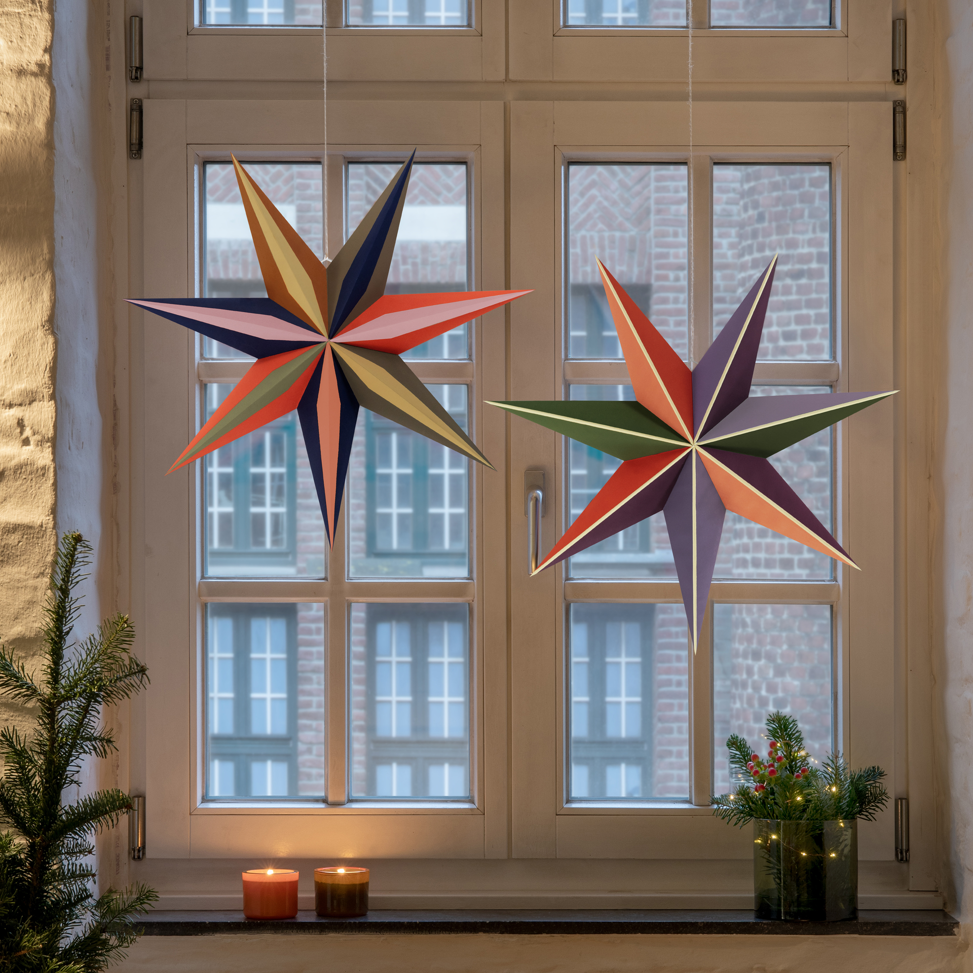 Deux étoiles en papier 'Sirius' sont suspendues devant une fenêtre. Des bougies, des branches de sapin et une guirlande lumineuse décorent le rebord de la fenêtre.