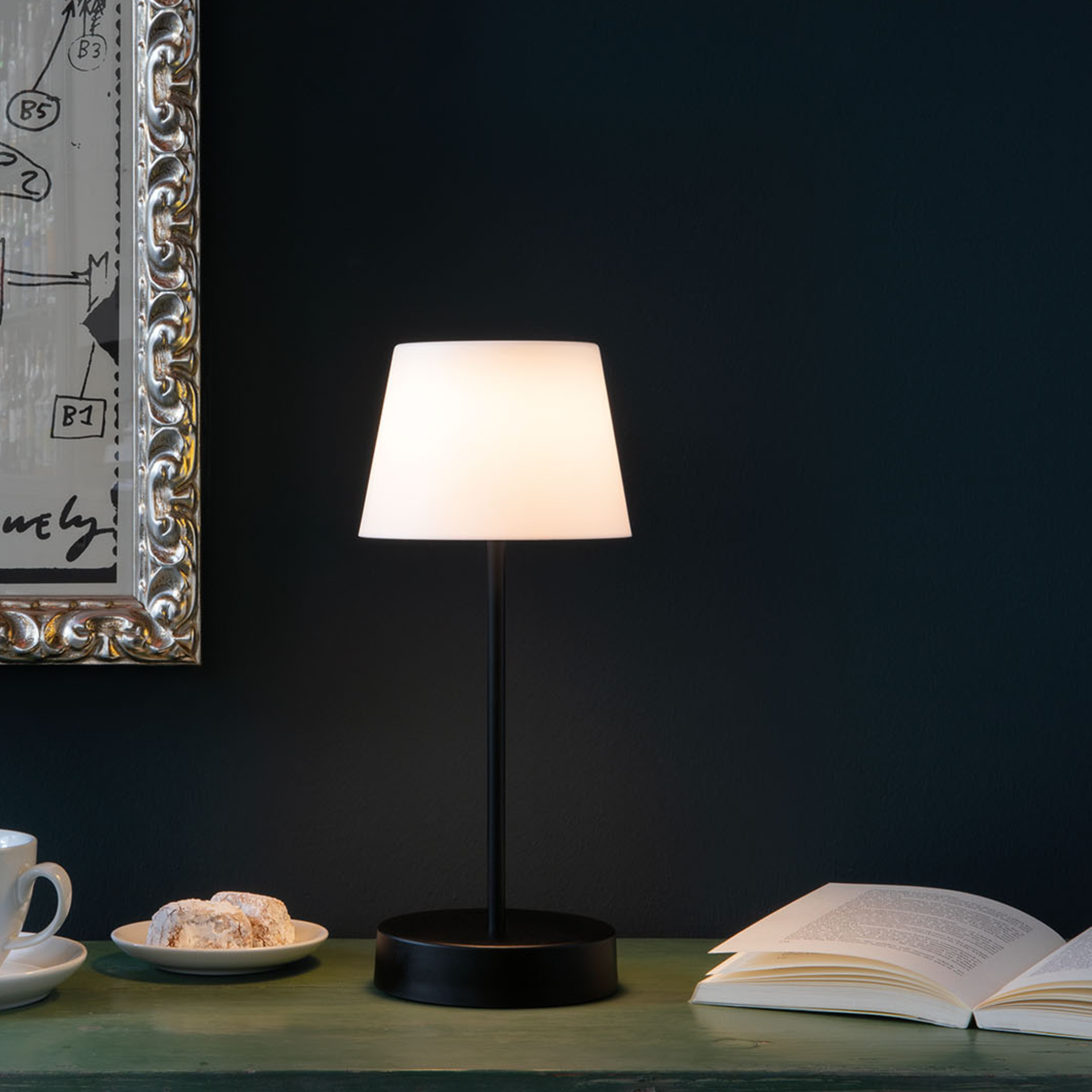 Tischleuchte Oscar 'pure' auf einem Tisch neben einem offenen Buch und einer Tasse. Die Leuchte hat einen schwarzen Fuß und eine weiße, leuchtende Kappe.