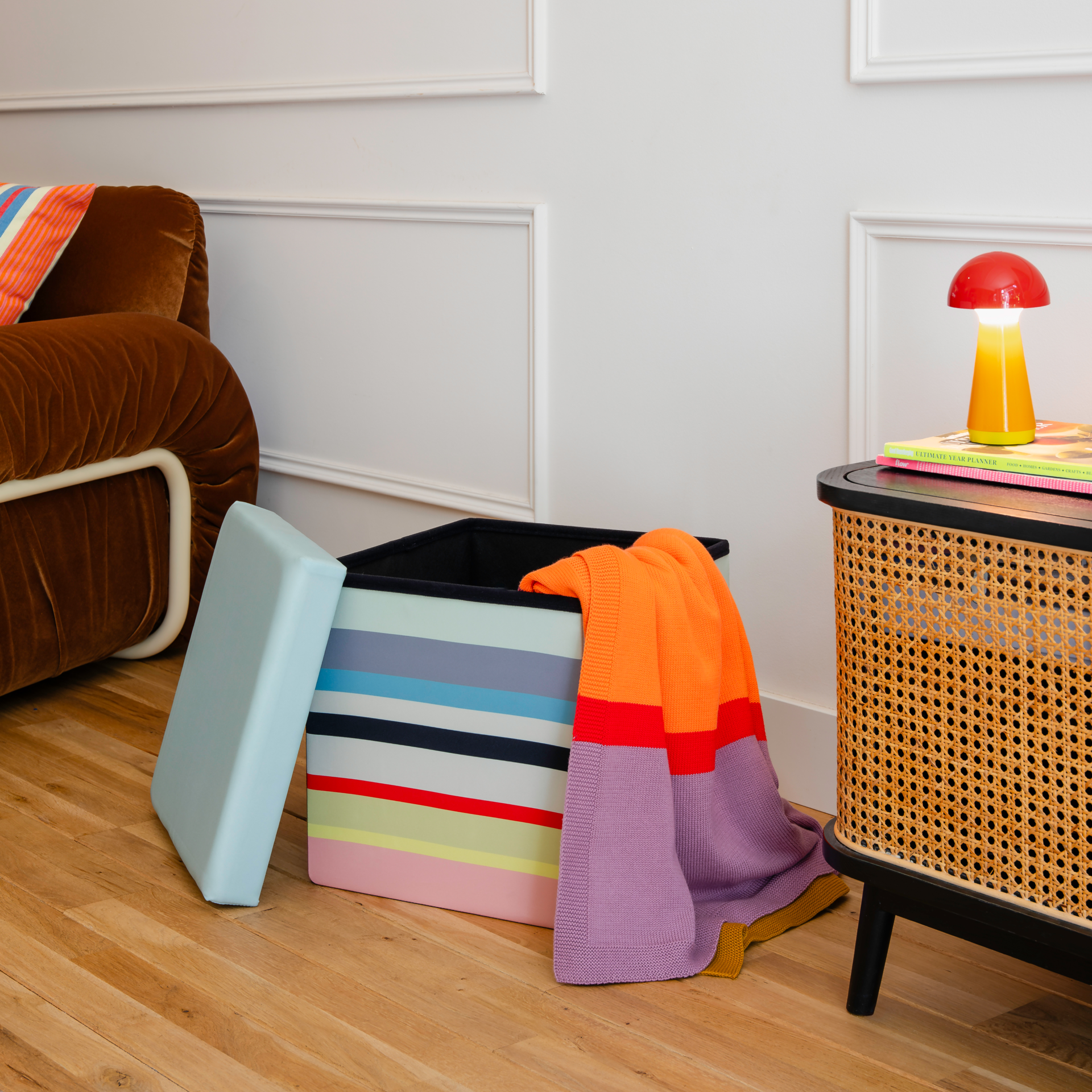 Boîte-siège cubique avec un couvercle vert clair. Côtés ornés de rayures horizontales dans les tons jaune pastel, gris, bleu clair, noir, rouge, jaune vif et pêche tendre, caractéristiques de la gamme « Remember 30 ». Siège et espace de rangement.