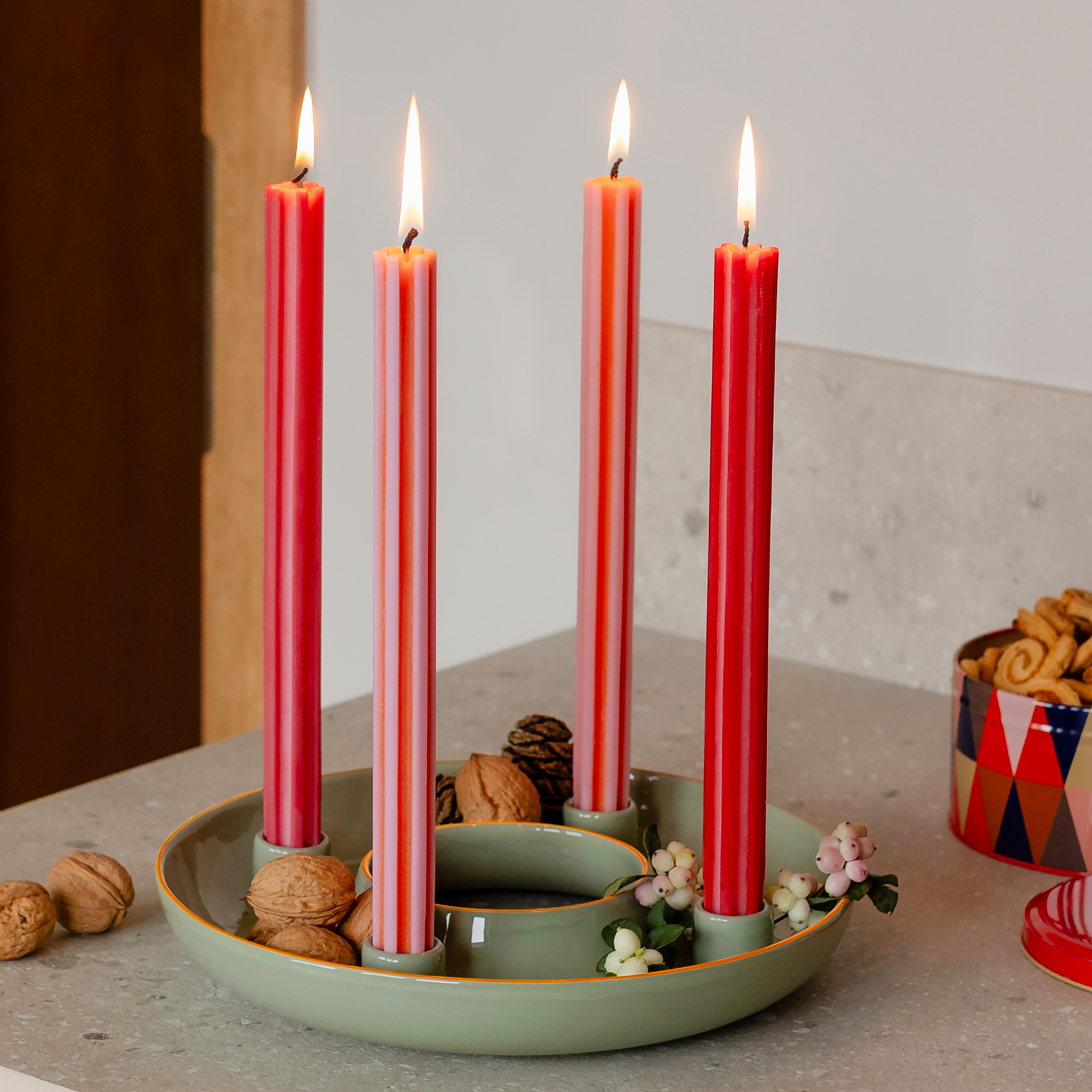 Ein Adventskranz mit vier brennenden Kerzen in Rot und rot-weiß gestreift, in einem grünen Kranz mit Walnüssen und weißen Beeren.
