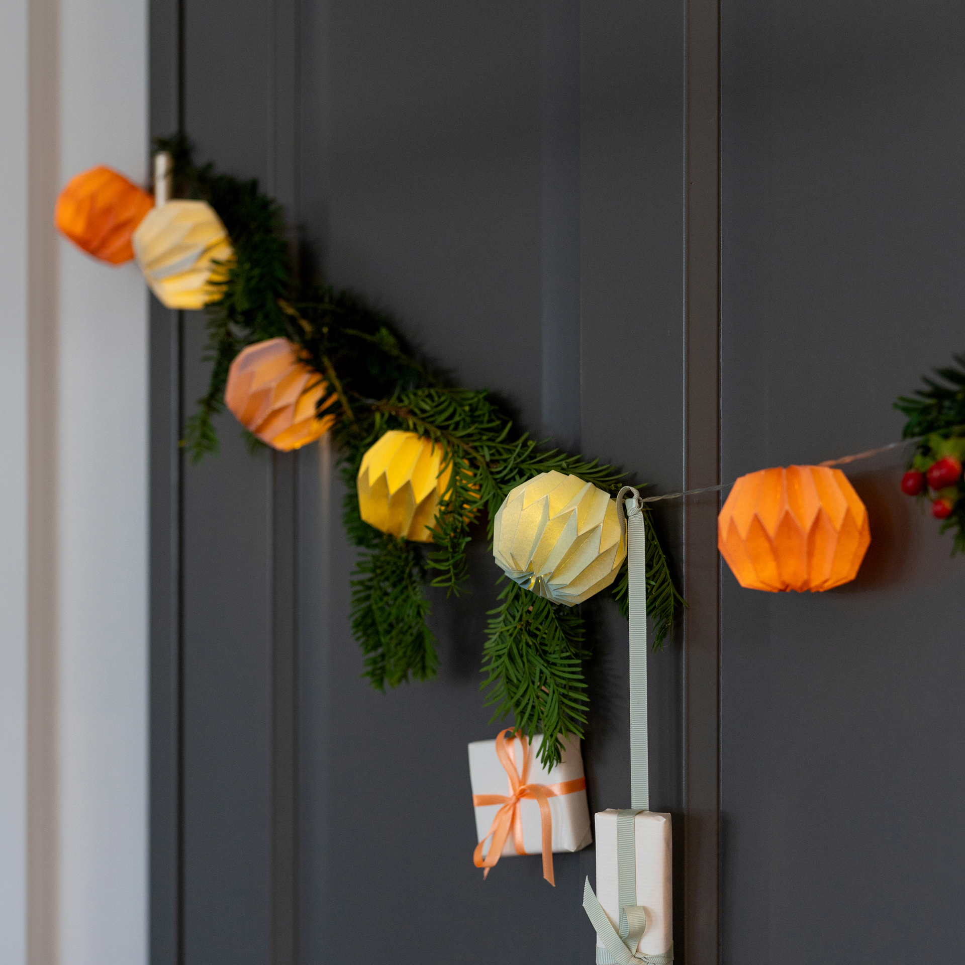 Eine Papier-Lichterkette Yazz, mit farbigen Papierornamenten und warmweißem Licht, dekoriert mit Tannengrün und kleinen Geschenken an einer grauen Wand.