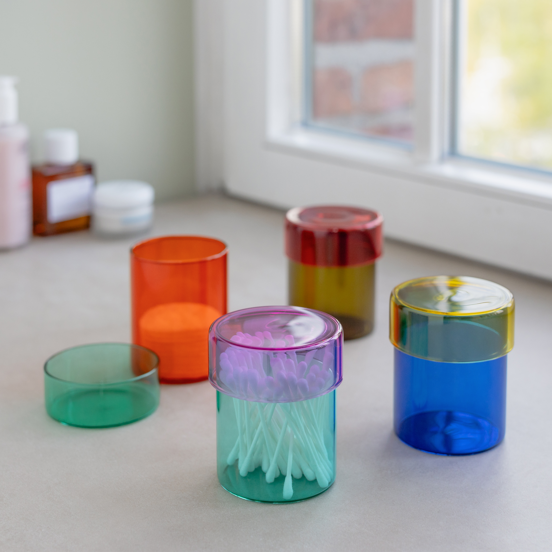 Fünf transparente Glasdosen mit Deckel in verschiedenen Farben, wobei eine mit Wattepads befüllt ist, sind auf einer hellen Oberfläche vor einem Fenster platziert.