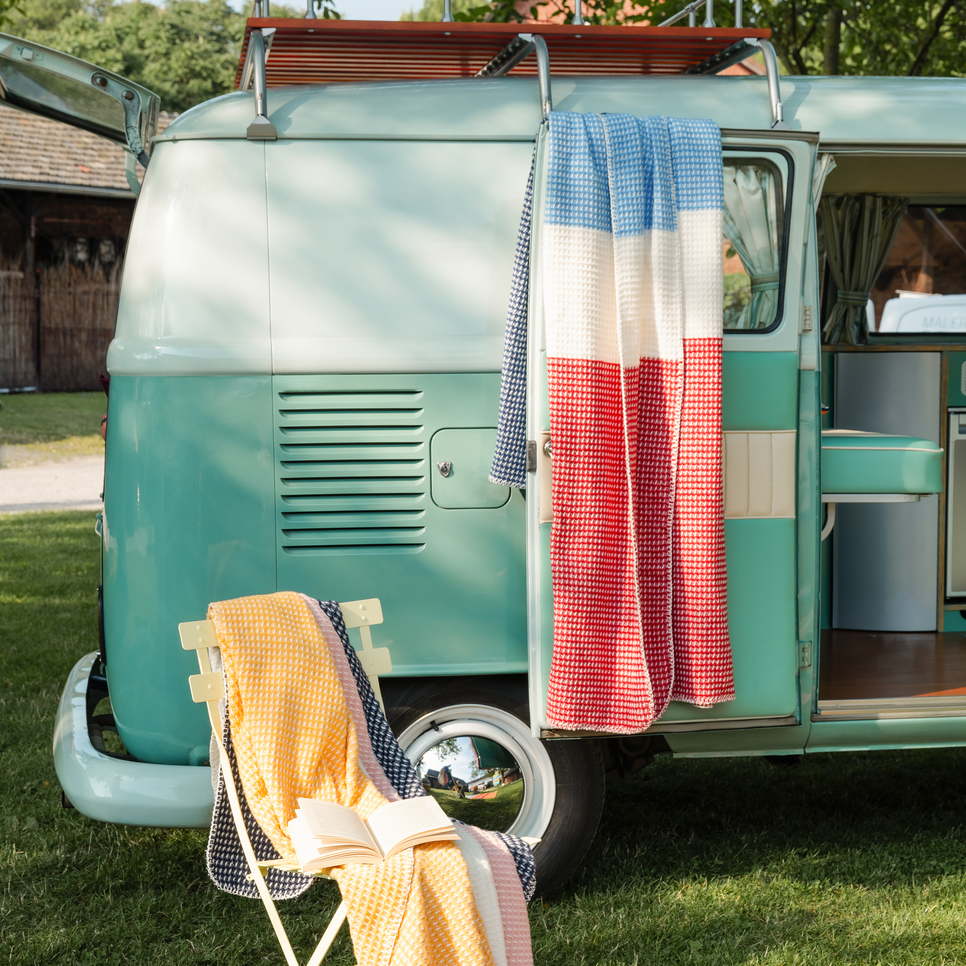 Ein hellblauer Campingbus mit offener Tür, darauf eine rot-weiß-blaue Decke. Auf einem Stuhl davor liegen weitere Decken und ein Buch.
