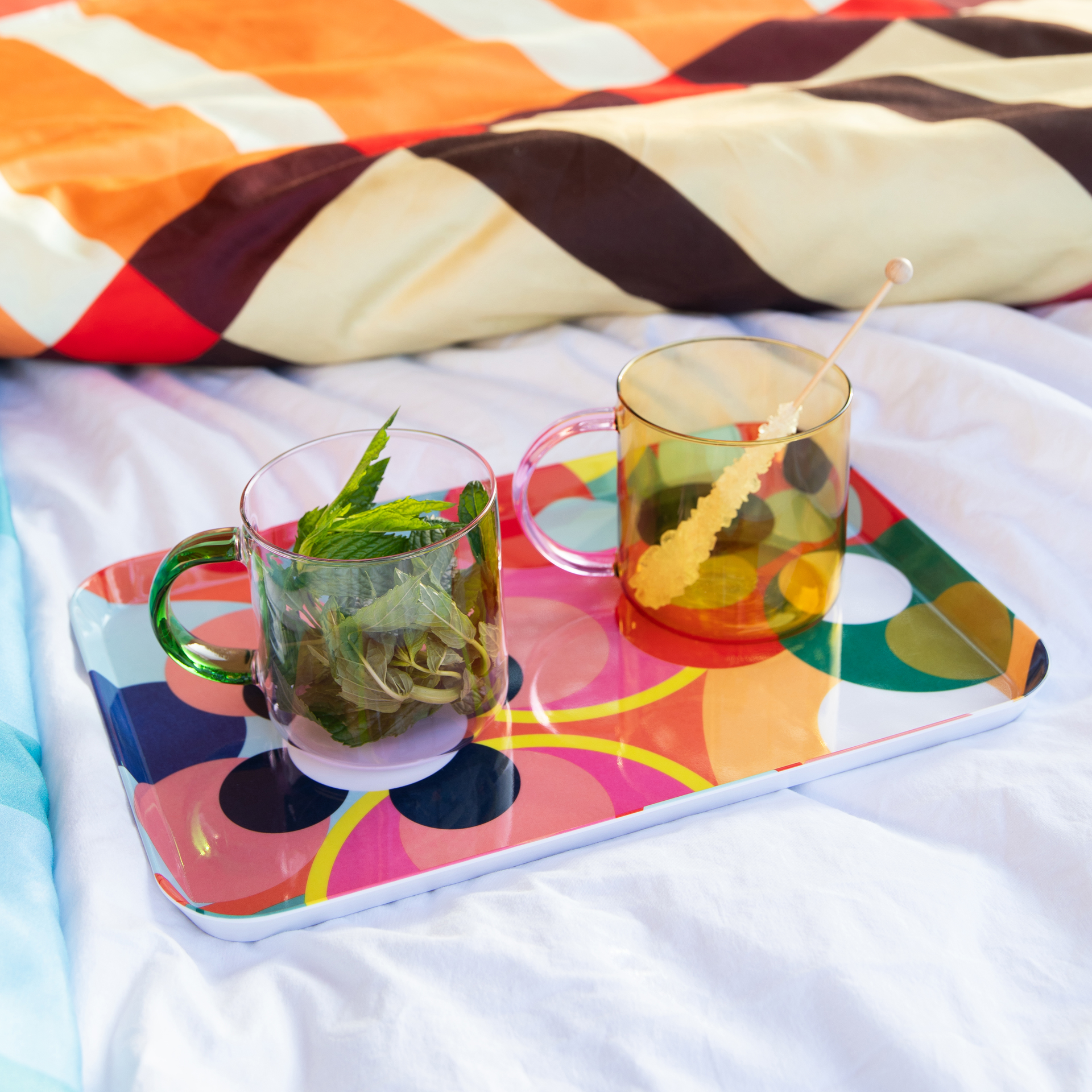 Zwei Glasbecher 'Fred' auf einem farbenfrohen Tablett im Bett. Der eine Becher mit Minze, der andere mit Kandis-Zucker am Stiel.