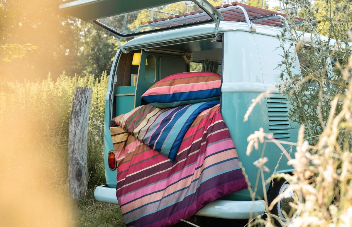 Un campercino azzurro, con il portellone aperto, è parcheggiato in un campo soleggiato e verde. Nella parte posteriore ci sono una coperta e cuscini a strisce colorate rosa, blu e terra, pronti per la notte.