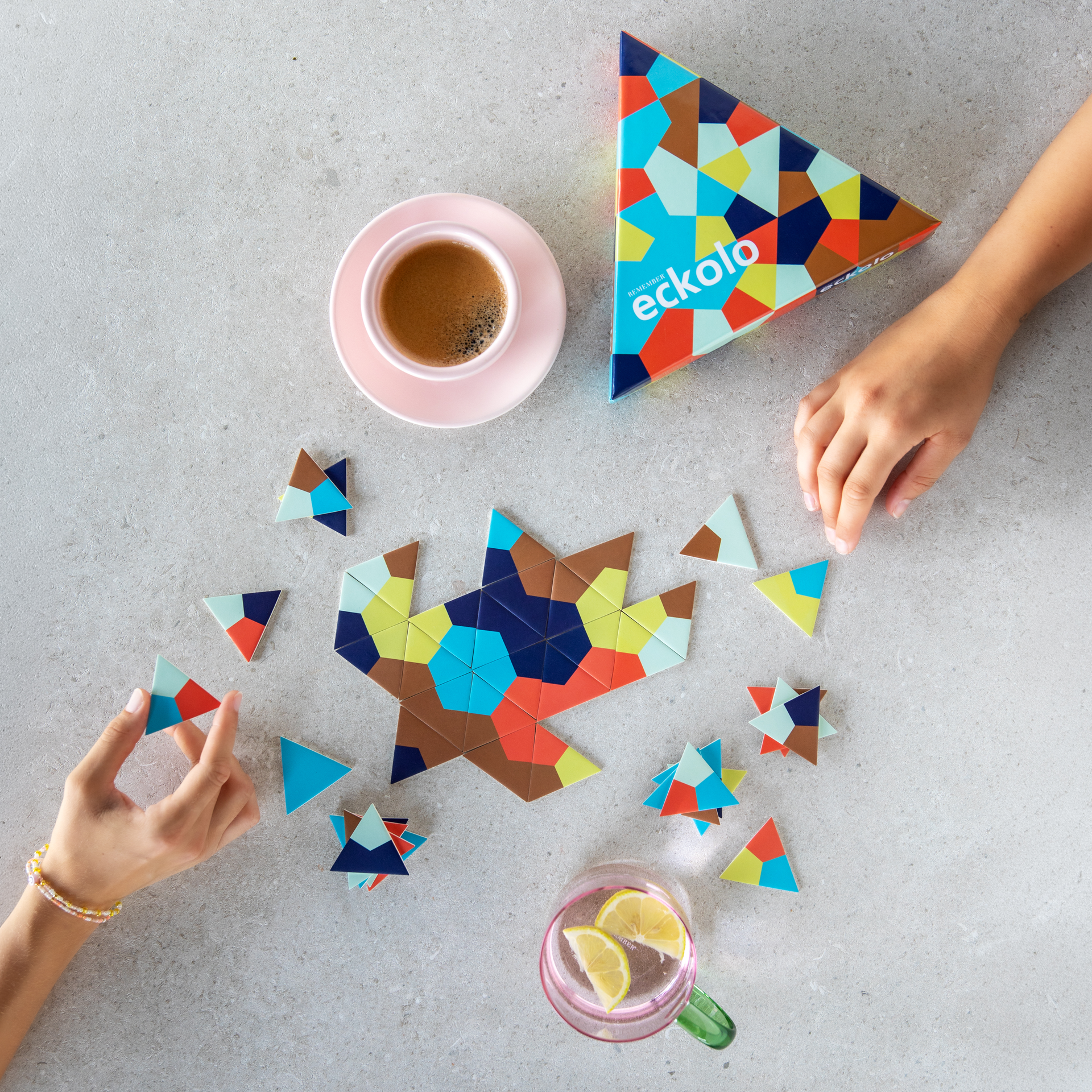 Boîte triangulaire du jeu Eckolo, ornée d’un motif géométrique coloré composé de polygones bleus, orange, jaunes et marron. Le logo « eckolo » apparaît deux fois, légèrement incliné, vu d’en haut sur fond blanc.