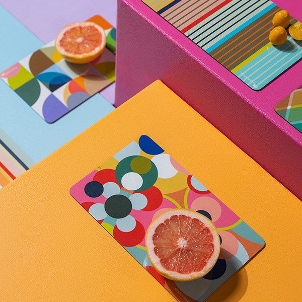 Bunte Tischsets mit geometrischen Mustern und Zitrusfrüchten auf farbigen Blöcken. Helle Farben dominieren die Szene.