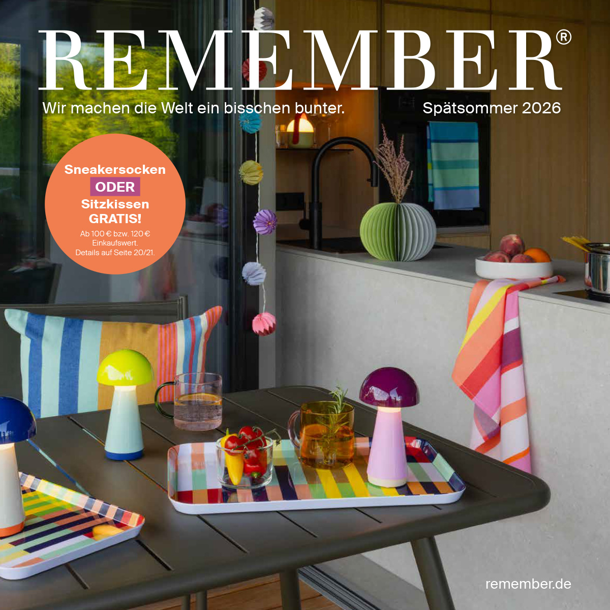 Terrasse élégante avec table foncée : lampes champignons colorées, boissons et en-cas sur plateaux rayés. En arrière-plan, cuisine moderne avec corbeille à fruits, torchon rayé et vase pliable vert. Guirlandes colorées.