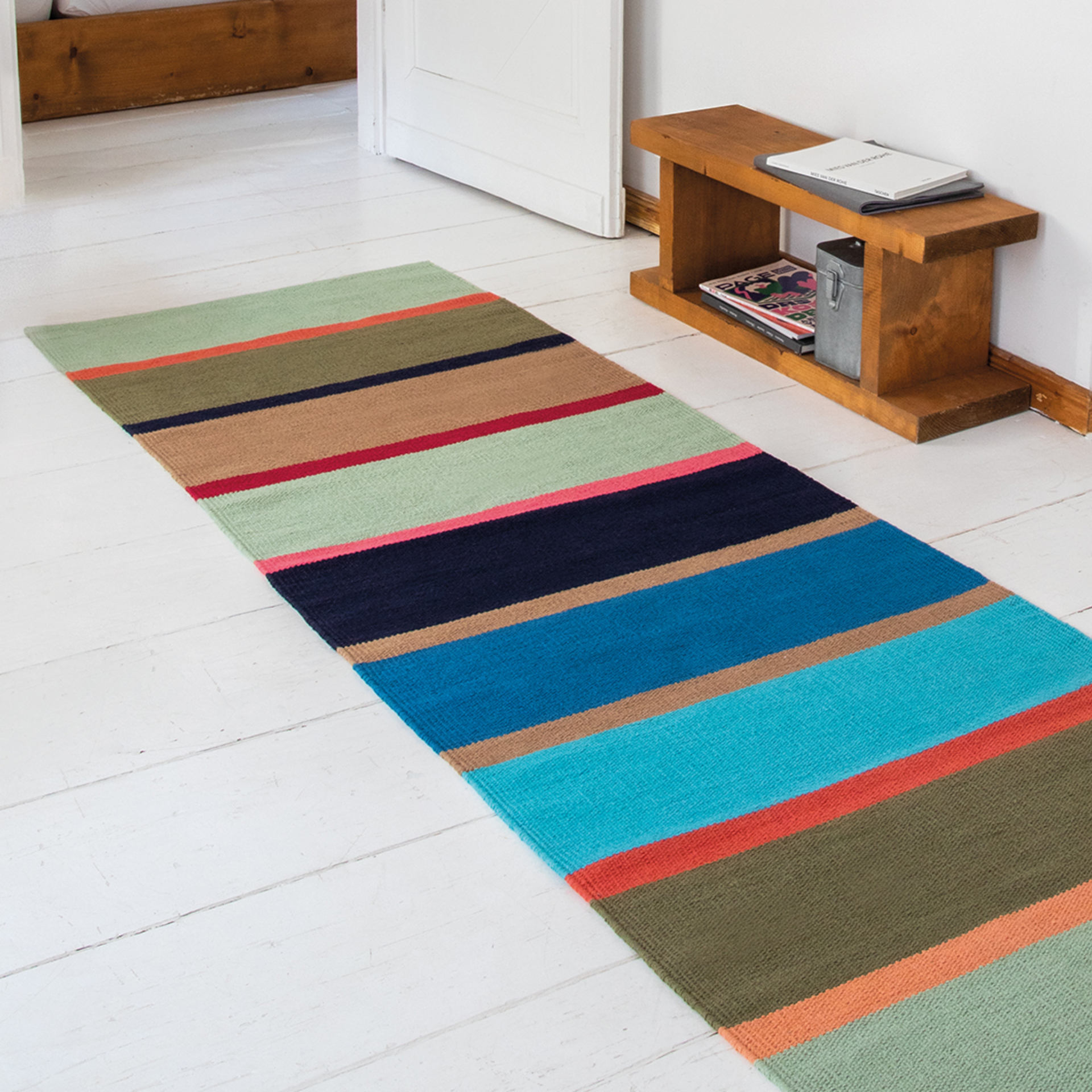 Langgestreifter Teppichläufer 'Costa' aus Baumwolle auf weißem Holzboden. Farben: Grün, Braun, Blau, Rot. Im Hintergrund ein Holzregal und eine offene weiße Tür.