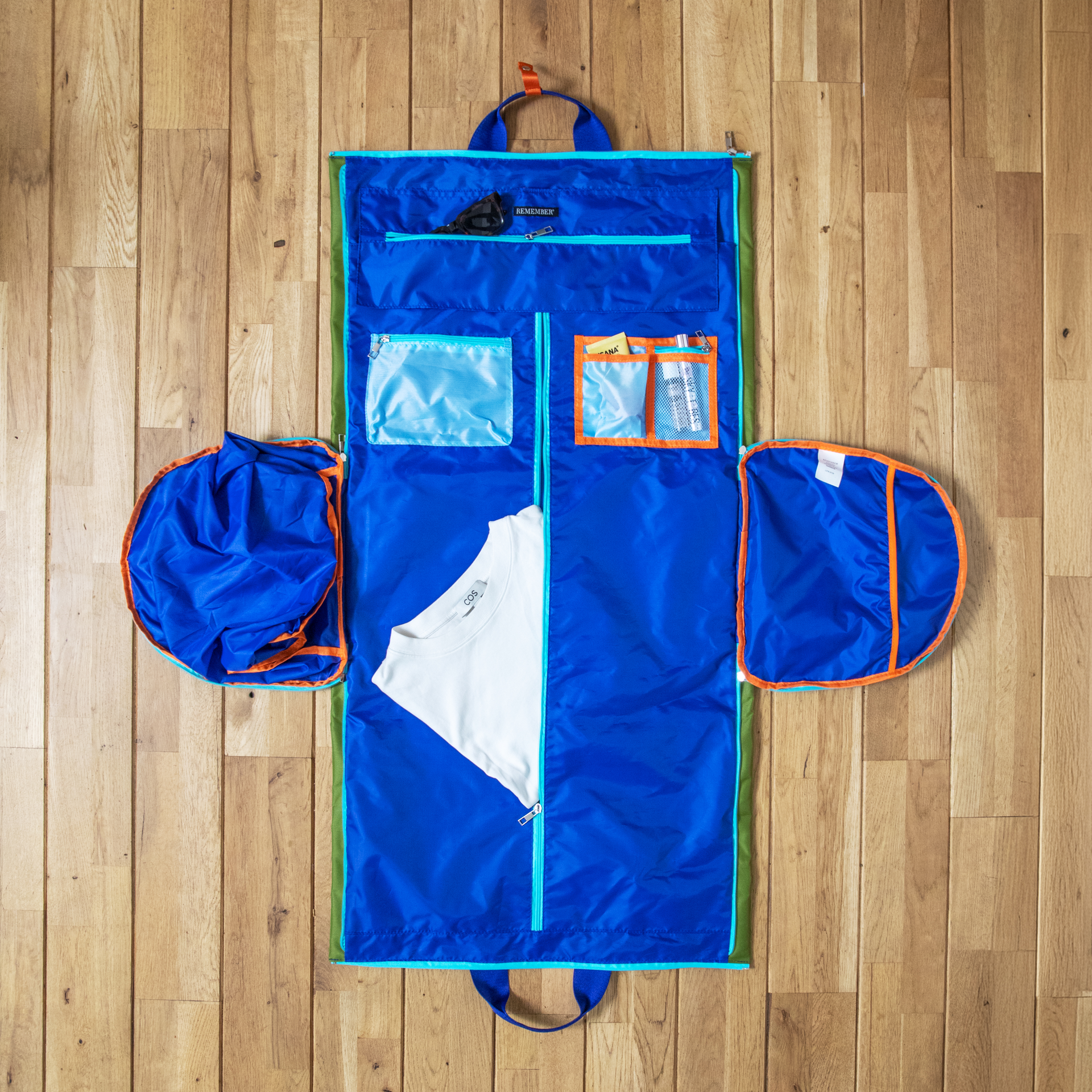Grand sac de voyage bleu Lennard, ouvert sur sol en bois clair. À l’intérieur : t-shirt blanc, lunettes de soleil, articles de toilette dans des compartiments. Compartiments latéraux ronds pour chaussures, dépliés.
